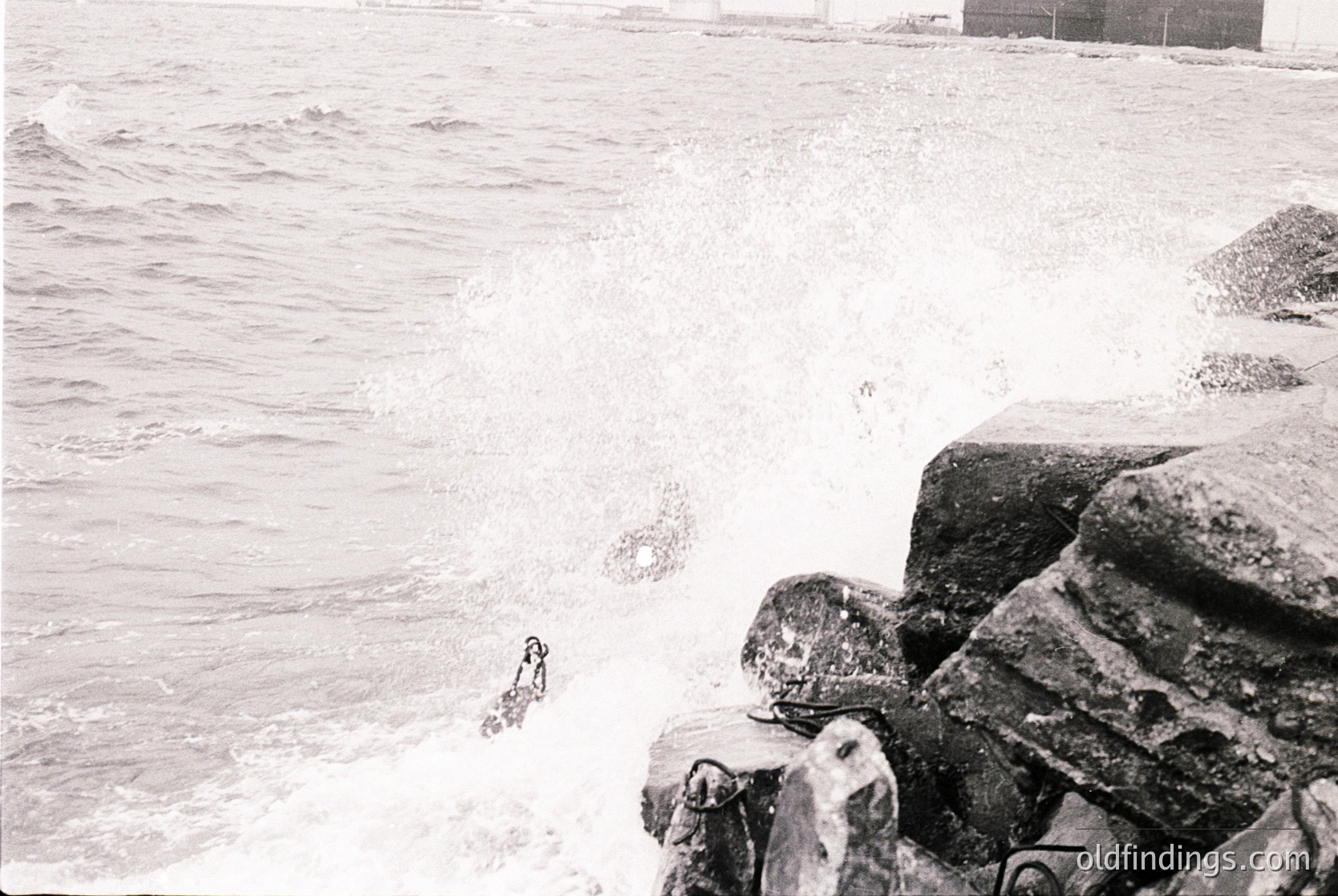 Black-and-white coastal scene featuring rough waves crashing against jagged rocks. A lone figure in mid-dive enters the water from the right, framed by the turbulent sea and rocky shore. Mid-20th century beach culture, likely or . Ideal for vintage travel, surf, or historical research.