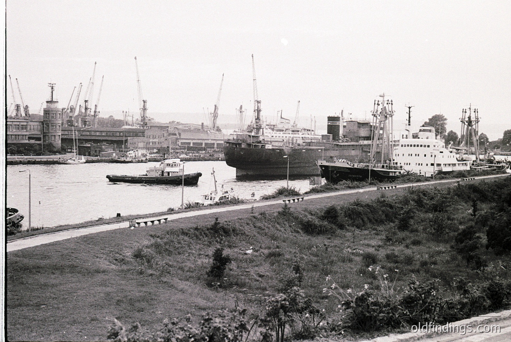 Black-and-white port scene featuring mid-20th century industrial maritime activity. Large cargo ships docked alongside cranes and warehouses, with smaller tugboats assisting. Prominent industrial architecture and waterfront infrastructure suggest a bustling trade hub. Likely Eastern European, mid-1950s–1970s.