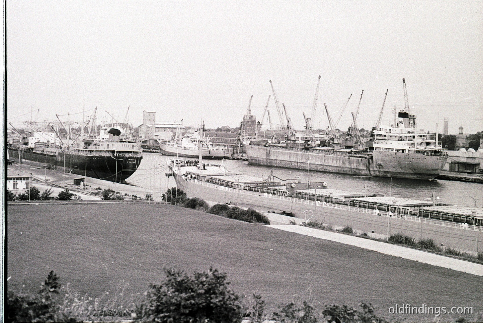 Black-and-white port scene featuring mid-20th century passenger ships docked alongside cranes and industrial infrastructure. Urban skyline with church spires visible in background. Likely Eastern European port, 1950s-1960s.