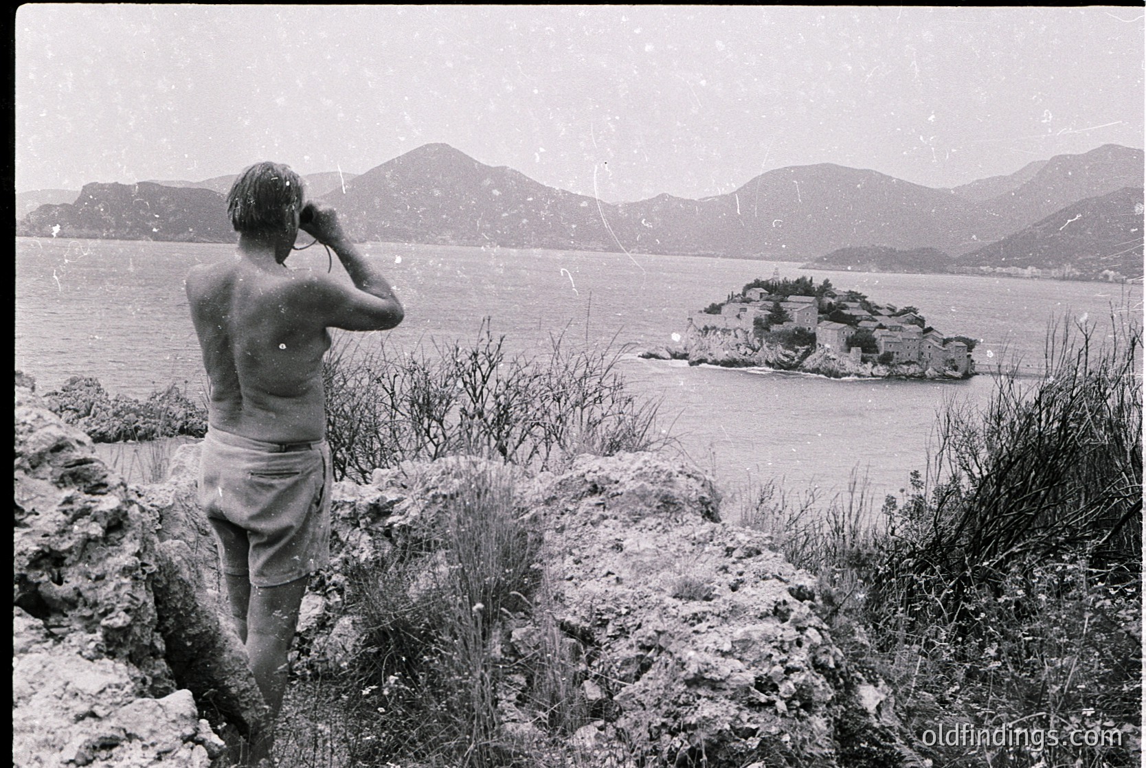A man in mid-20th-century swimwear poses on rocky terrain, facing a rugged island fortress in a coastal bay. Snow-capped mountains frame the scene, suggesting a Mediterranean or Black Sea locale. Classic black-and-white composition captures 1950s–60s seaside exploration.