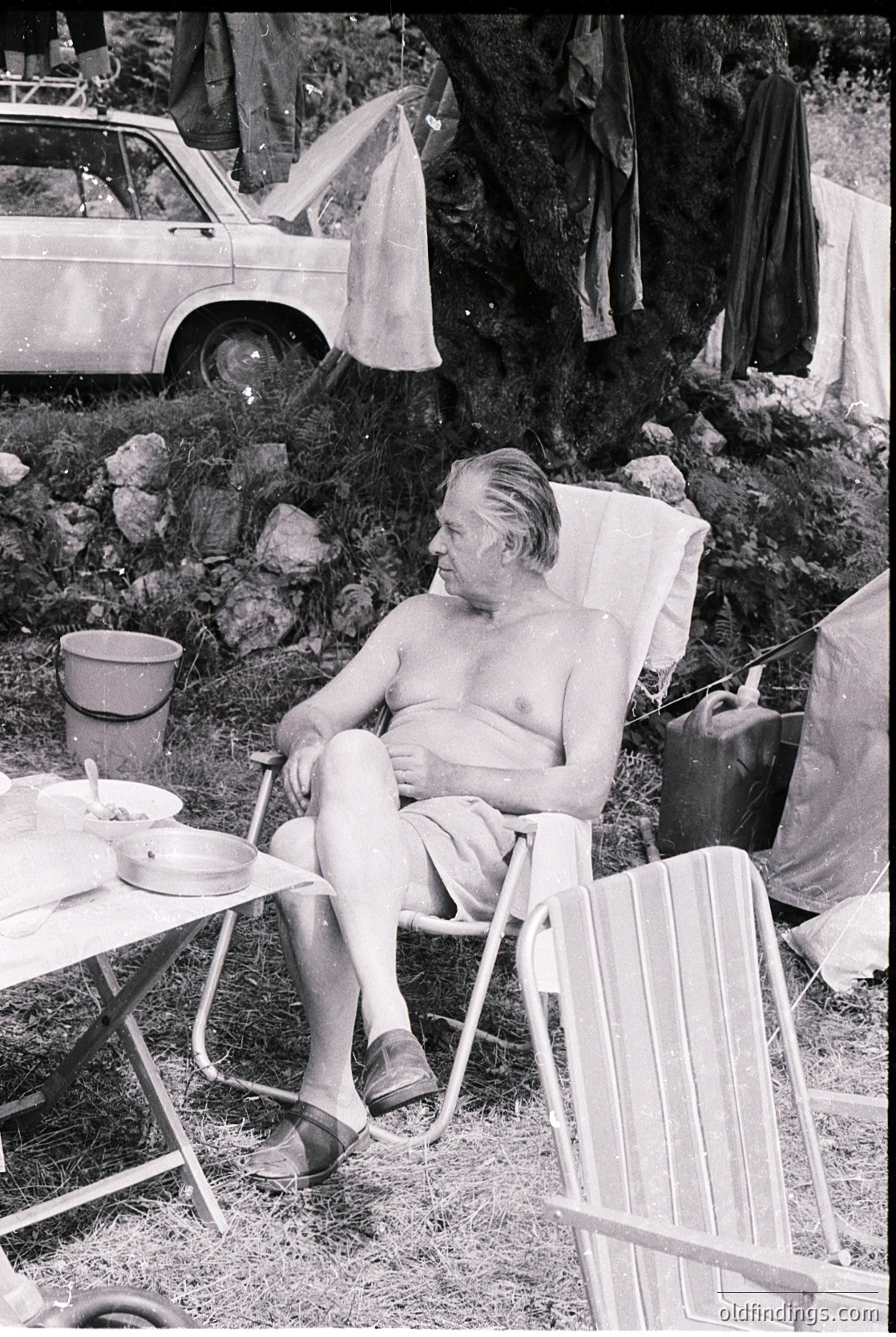 Mid-20th century man lounges in a vintage deck chair beside a vintage car and camping setup. Rock formations and a makeshift tent frame the scene, suggesting a rugged outdoor retreat.