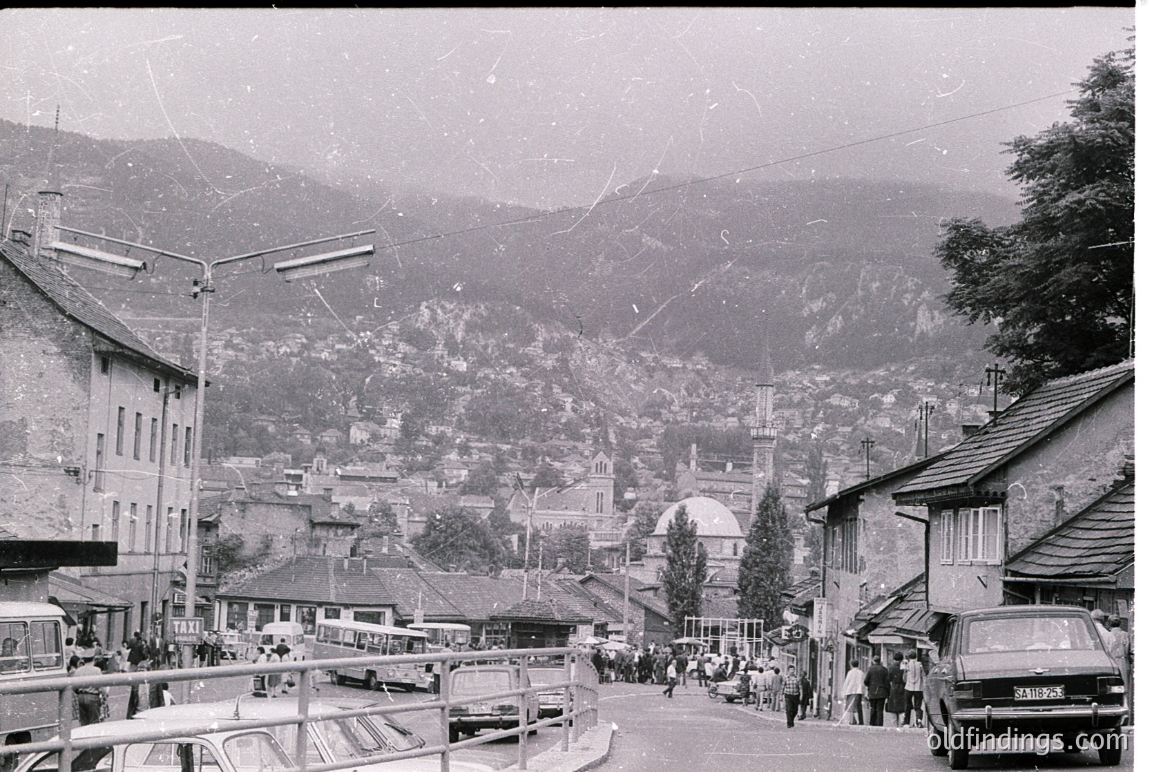 Vintage black-and-white street scene in a mountainous European town, likely 1950s–1970s. Wooden buildings with sloped roofs line a bustling pedestrian area, flanked by parked cars and a tram line. A mosque with domes and minarets stands prominently in the background, suggesting a blend of Alpine and Ottoman architectural influences. Crowds gather near market stalls, hinting at a lively local economy.