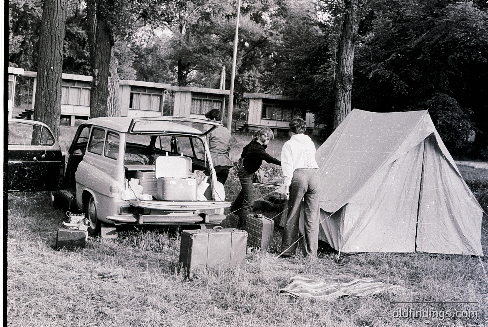 Mid-20th century camping scene: two men setting up a canvas tent near a vintage station wagon loaded with luggage and a cooler. Surrounding wooden cabins suggest a campground or resort. Black-and-white, likely 1960s–1970s.