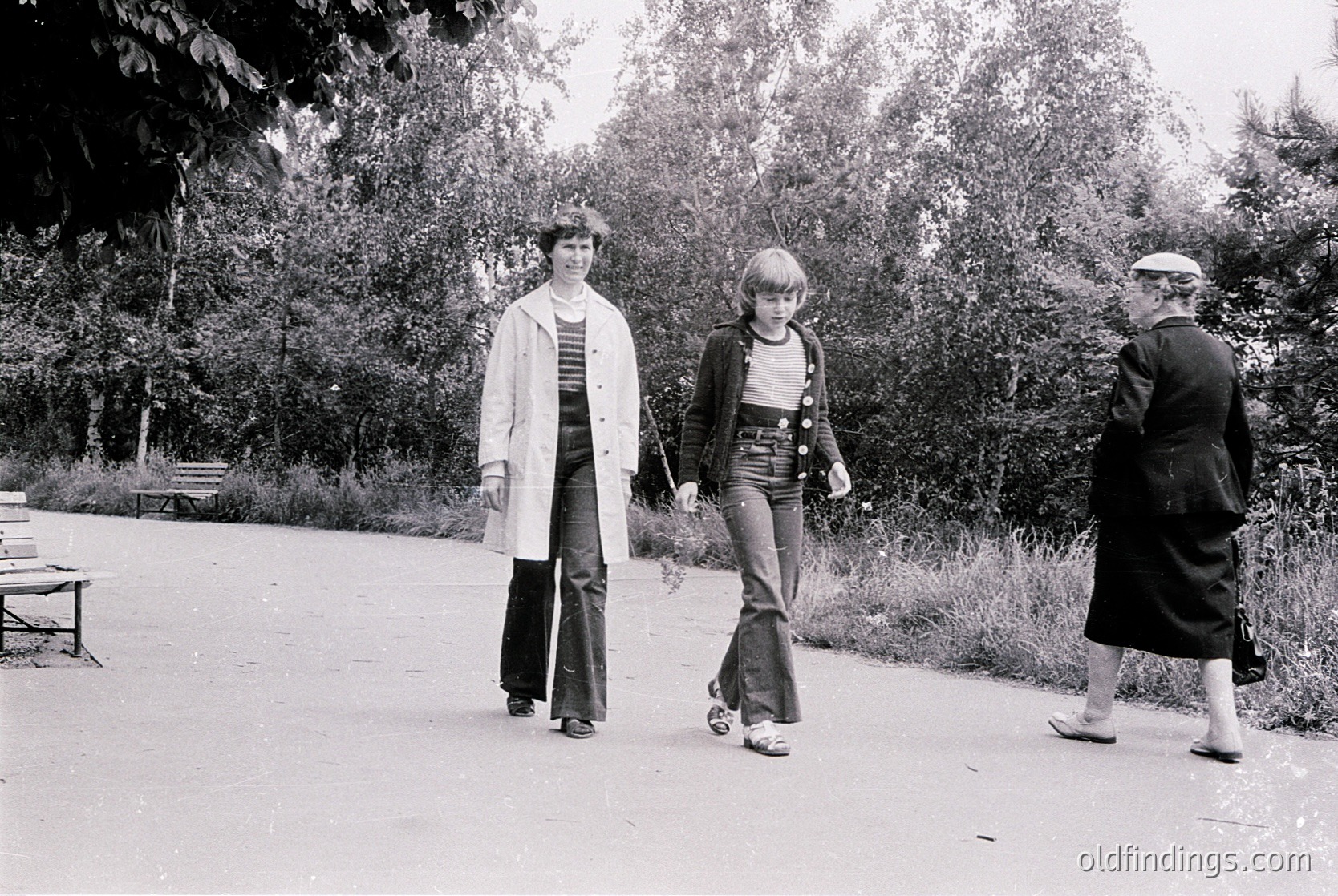 Three individuals stroll along a tree-lined park path in 1970s fashion: flared jeans, striped tops, and layered cardigans. Lush greenery and a wooden bench frame the scene, suggesting a suburban or urban park setting. Candid, black-and-white snapshot captures youthful leisure.