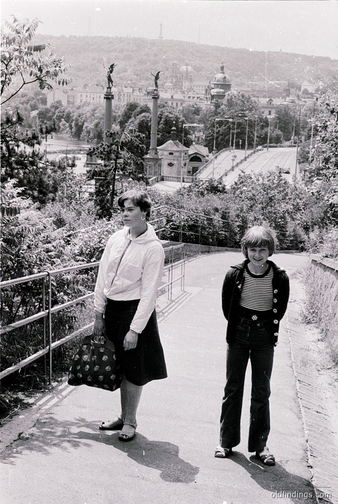 Two individuals pose on a sunlit urban pathway, likely mid-20th century (1960s–70s). The woman wears a knee-length floral-patterned skirt, white blouse, and sandals; the man sports a striped sweater, dark trousers, and loafers. A vintage tram or trolley station and lush greenery frame the background, hinting at a European cityscape. The composition captures mid-century fashion and urban design.