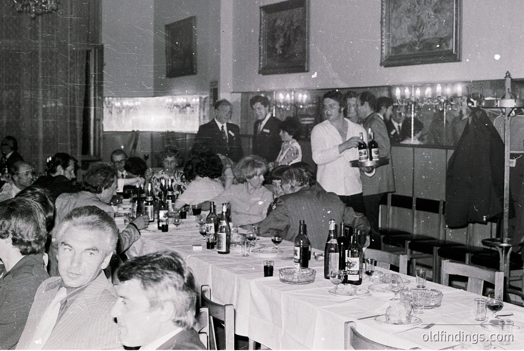 Formal banquet hall with long tables draped in white linen, featuring wine bottles, glasses, and place settings. Attendees in 1960s–70s formalwear—suits, dresses, and name tags—engage in conversation. A waiter in a white apron pours wine. Ornate chandeliers and framed artwork adorn the walls, suggesting an upscale venue.
