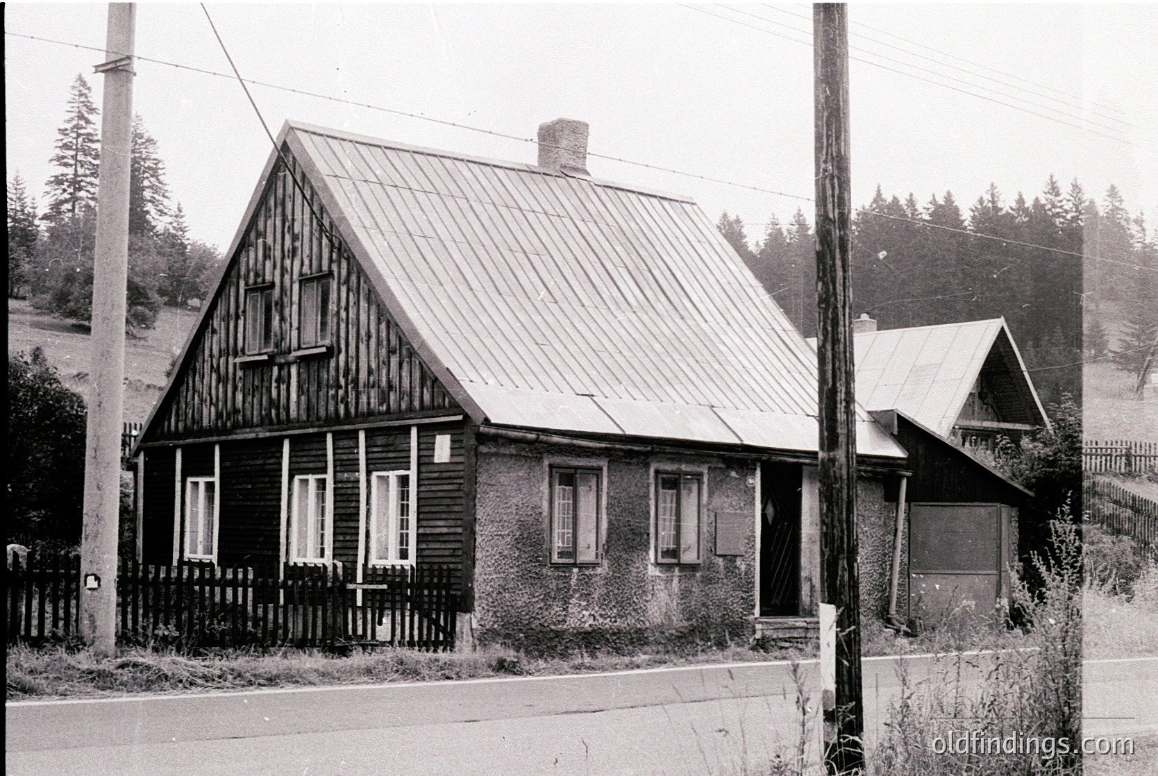 Mid-20th century wooden house with gabled roof and white-framed windows, set in a rural forest edge. Brick chimney and picket fence detail visible. Black-and-white vintage photograph.