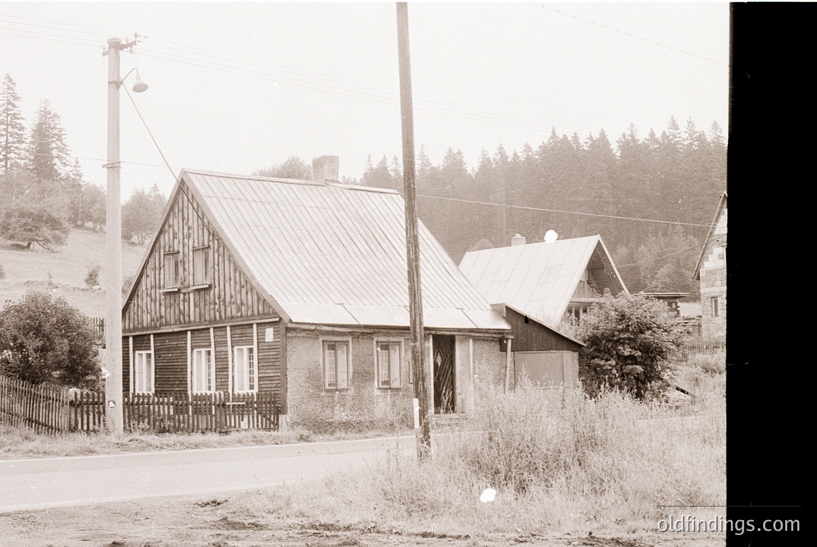 Rustic wooden houses with gabled roofs in a rural setting, likely early-to-mid 20th century. Wooden picket fence and overgrown vegetation suggest abandonment or minimal maintenance. Utility poles and wires indicate electrification.