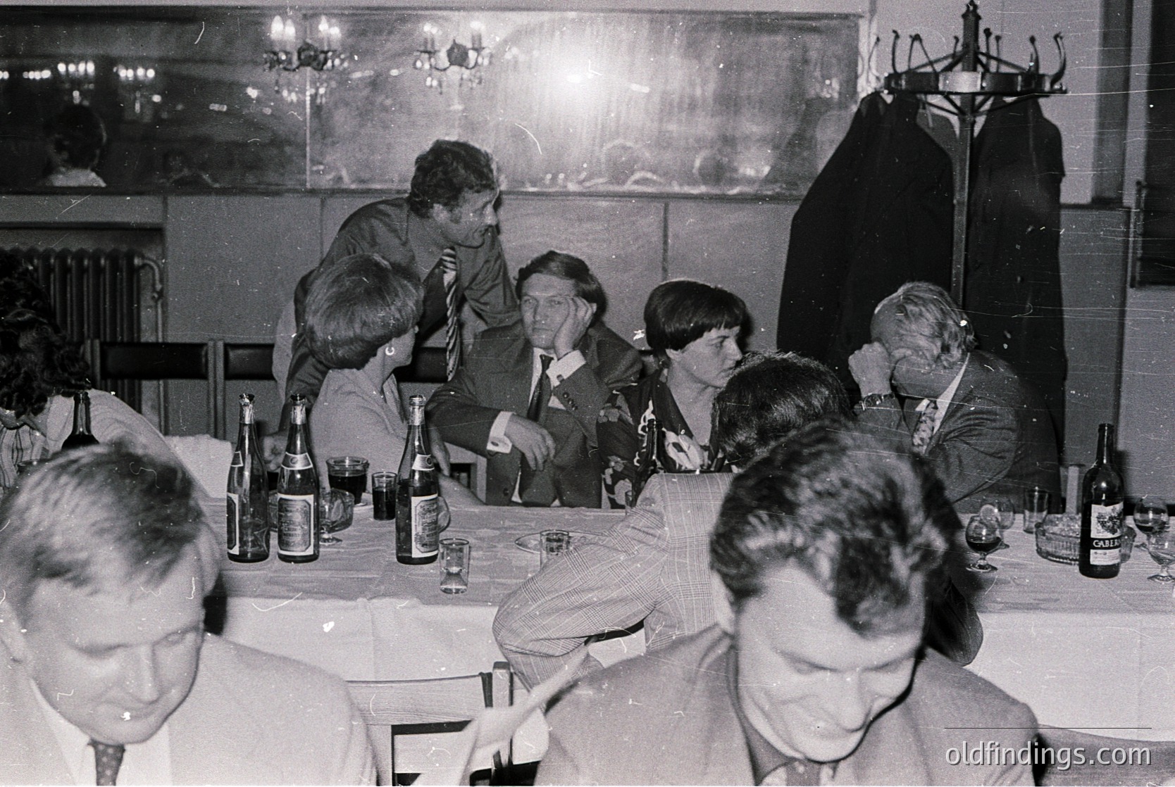 Black-and-white gathering of men in formal attire at a mid-20th century banquet, likely 1960s–1970s. Bottles of *Balkan* beer and glasses on a long table with chandelier lighting. Indoor setting with draped curtains and coat hooks. Social event with relaxed postures and casual conversation.