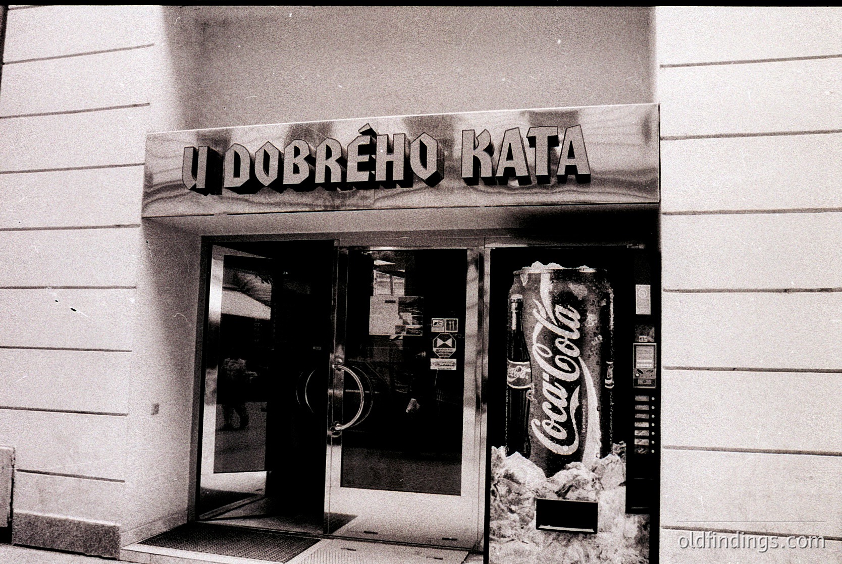 Vintage storefront with Cyrillic sign "У Добрехо Ката" (likely Bulgarian for "Good Cat") above double glass doors. Coca-Cola vending machine on right, snow piled below. Mid-century commercial architecture, urban setting.