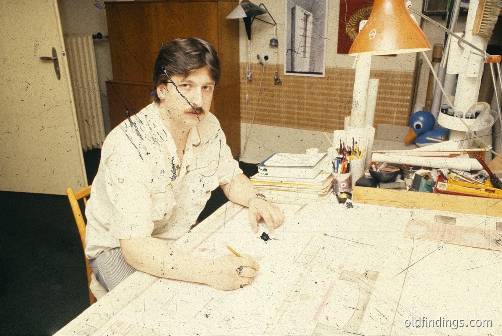 Mid-century artist engrossed in abstract painting, seated at a cluttered wooden table with paint-splattered clothing. Surrounding materials include a lamp, sketchbooks, and a duck-shaped desk organizer. Likely 1960s–1970s, Western industrial setting.