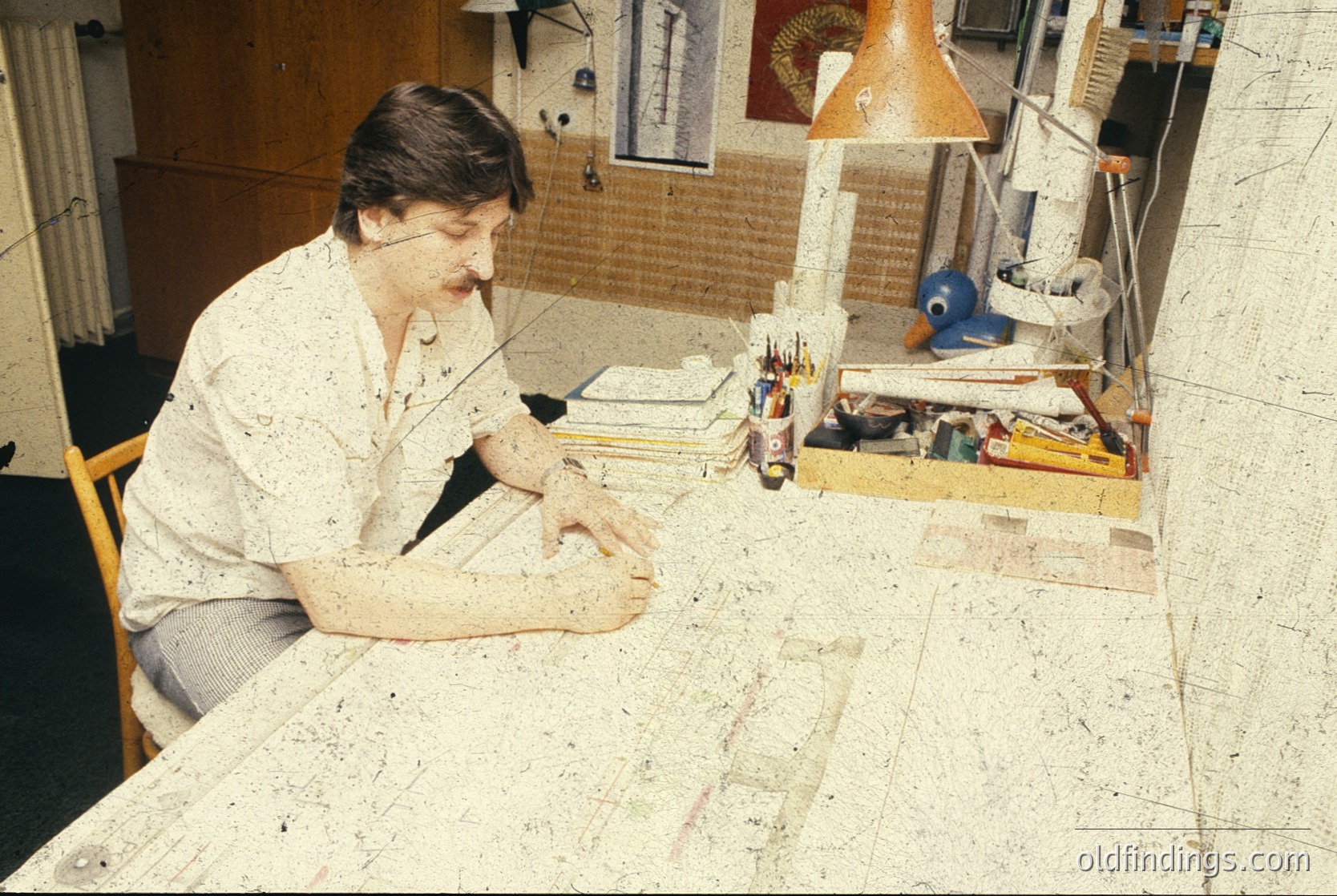Mid-century workspace featuring a focused individual engrossed in detailed work on a large, flat surface. Surrounding materials include stacked books, a lamp with a beige shade, and a wooden crate filled with art supplies. The setting suggests a creative or technical environment, likely from the **1960s–1980s**.