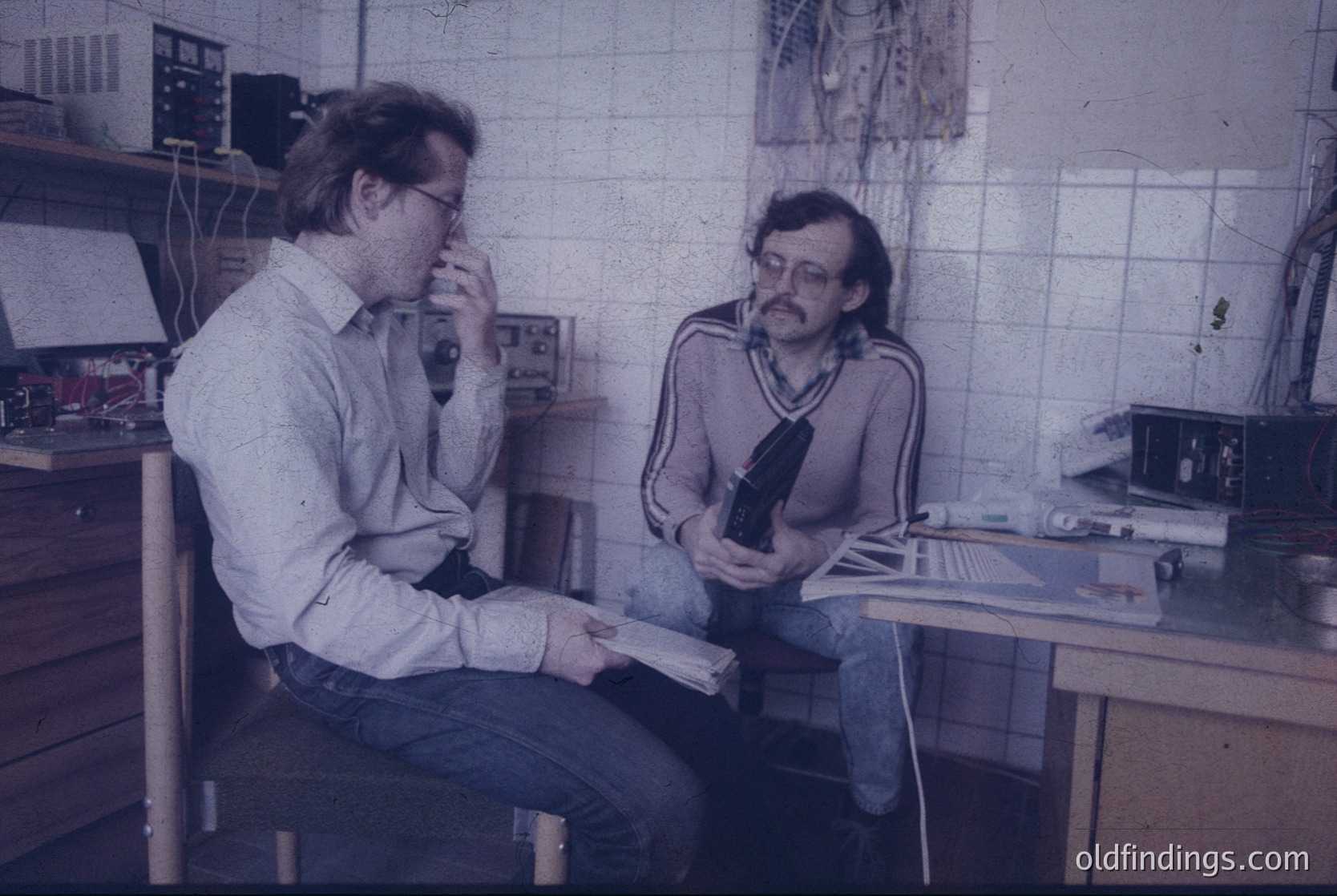 Two men in a 1970s-era office setting, likely Eastern Bloc. Left: man in a button-down shirt, holding a rolled document, seated on a stool. Right: man in a striped sweater, examining a camera with a long lens. White-tiled walls, basic wooden furniture, and analog equipment (typewriter, tape recorder) suggest a mid-century workspace. Potential historical research or industrial design reference.