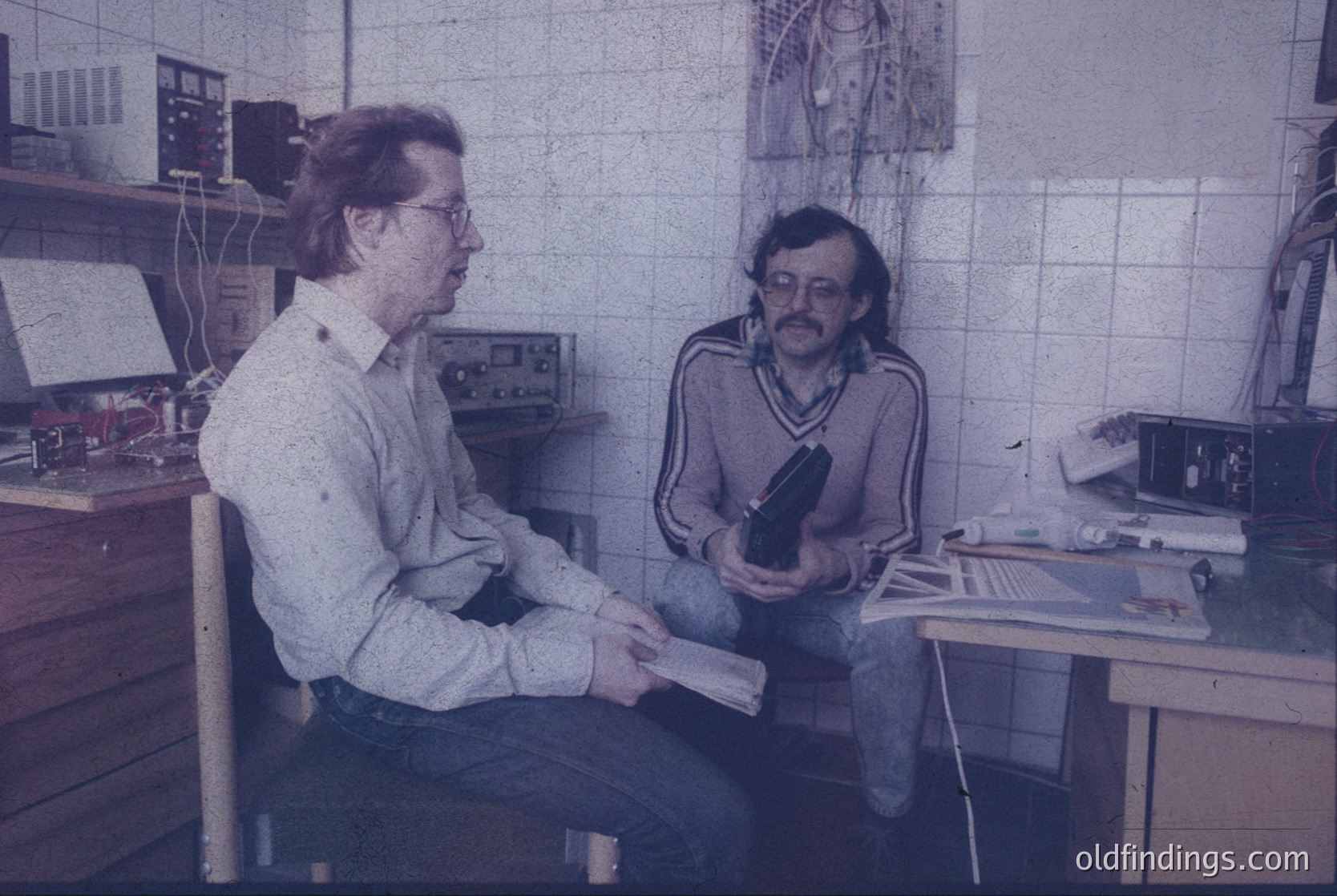 Two men in a retro office setting, likely mid-1970s–1980s. One holds a camera with a large lens, the other examines printed photos. White-tiled walls, analog equipment (typewriter, desk lamps), and a framed poster in the background suggest a photography studio or lab.
