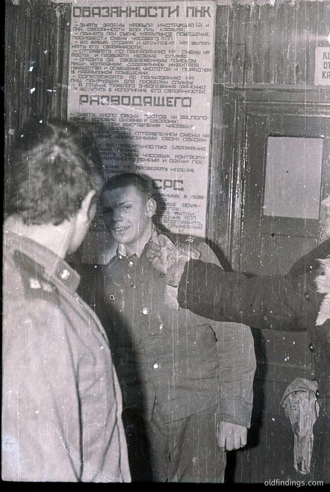 Two men in Soviet-era uniforms stand in front of a wooden door with Cyrillic text. The man on the right points toward the door while smiling, likely at a checkpoint or entrance. The signage references "obligations" and "duties" ( ).