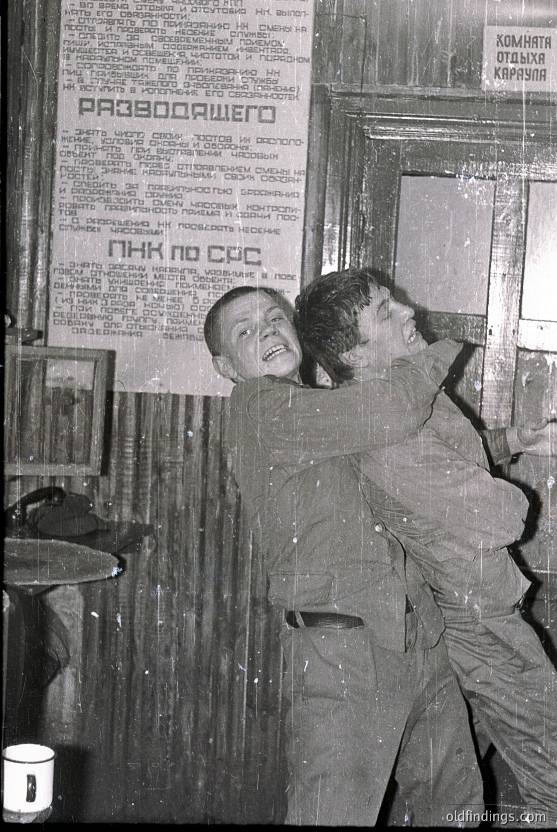 Black-and-white photo of two men in heavy work uniforms (likely miners) leaning against a wall with Cyrillic text—likely Bulgarian—posted above them. The sign advertises a holiday resort ("Комната отдиха Караван"). Mid-20th century Eastern Bloc industrial setting.