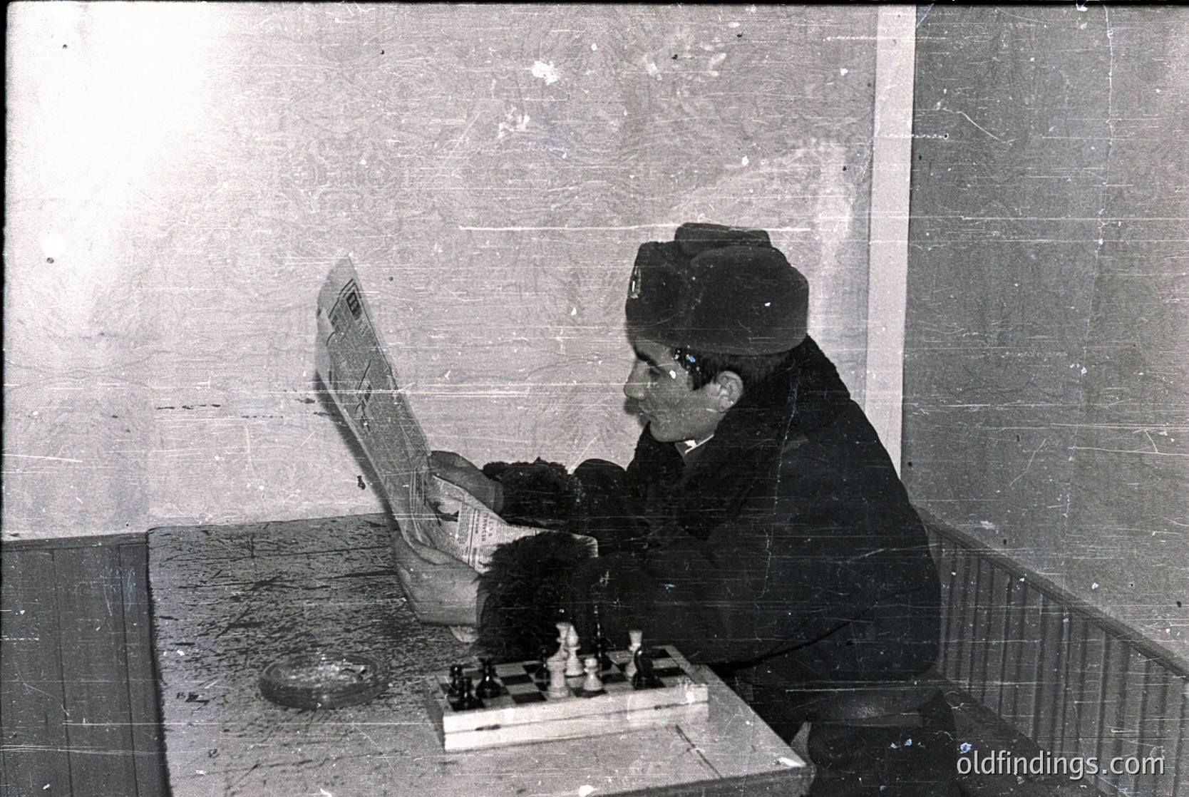 Vintage black-and-white photo of an elderly man in a peaked cap and heavy coat playing chess in an indoor setting, likely a public space. Chessboard and pieces in focus on a wooden table. Mid-20th century attire suggests or .