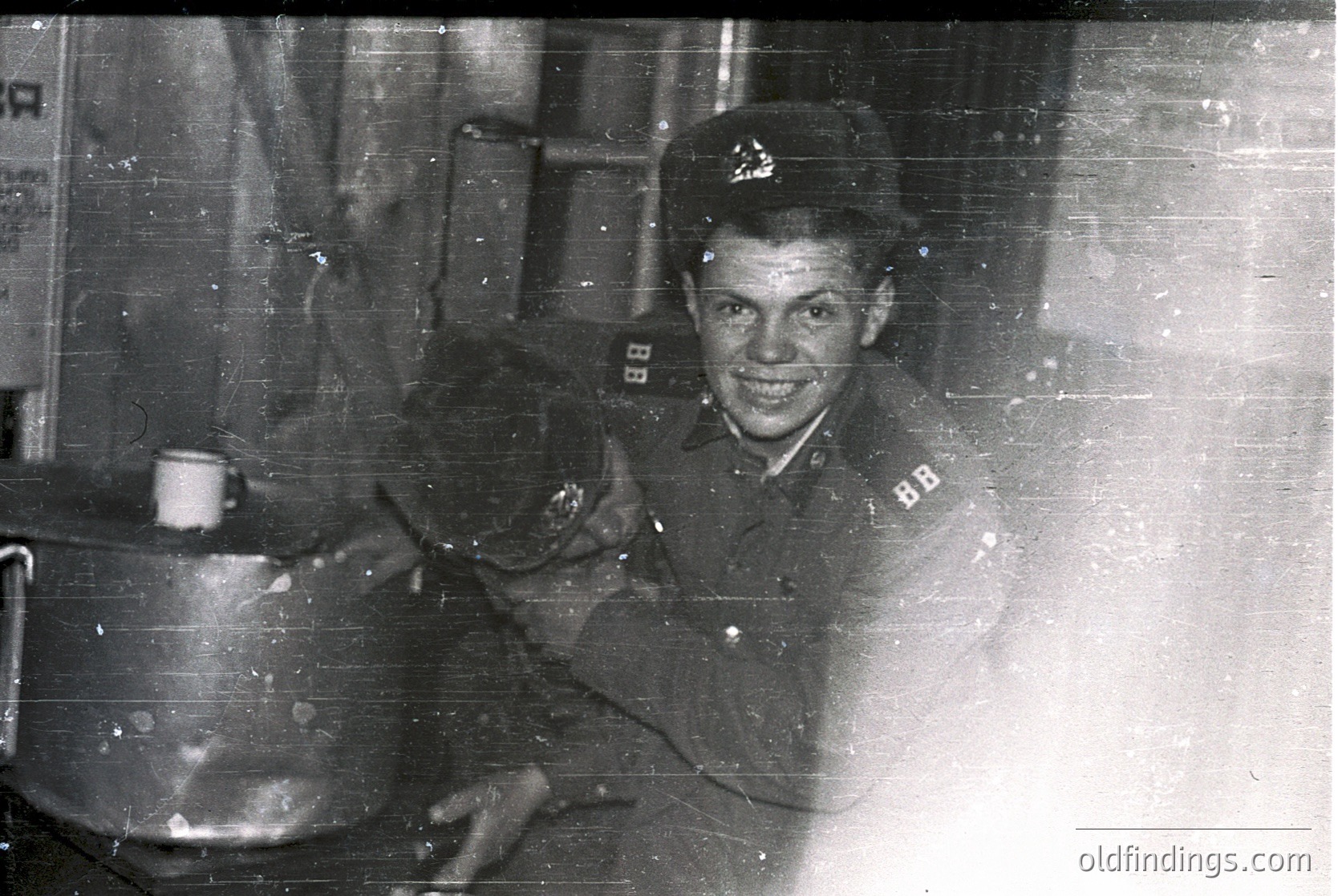 Young man in mid-20th century military uniform, likely Soviet-era, holding a large pot. Indoor setting with visible industrial or domestic equipment. Uniform patches suggest possible naval or transport corps affiliation. Vintage black-and-white photo with slight wear.