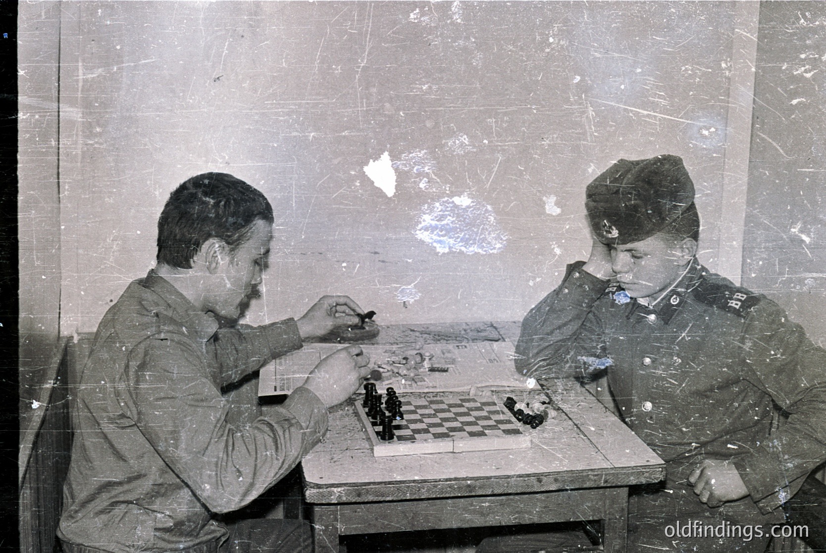 Two uniformed men play chess in a dimly lit, weathered room, likely mid-20th century. Uniforms suggest military or state service; one wears a peaked cap with insignia. Faded wall and table indicate aged or archival quality.