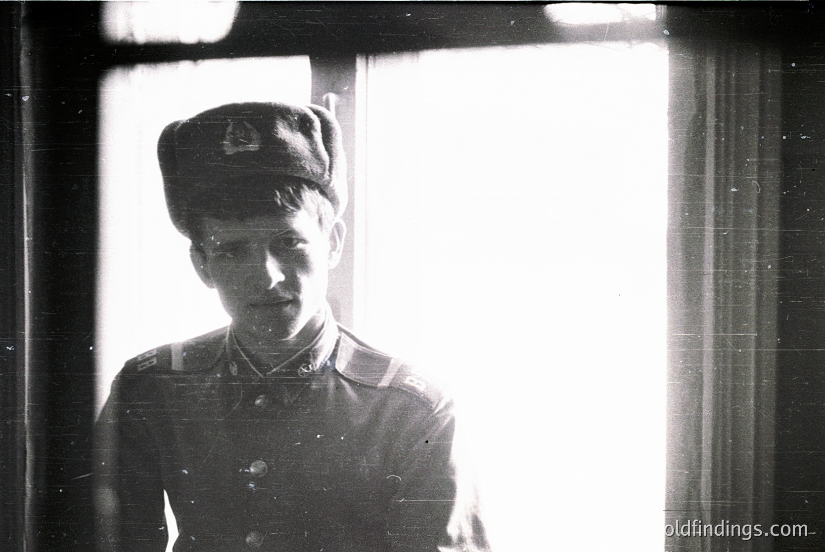Young man in early 20th-century military uniform, likely 1910s–1930s, standing indoors near a window. Uniform includes peaked cap with insignia, dark tunic with collar details, and a visible shoulder cord. Vintage black-and-white grain and lighting suggest candid or documentary style.