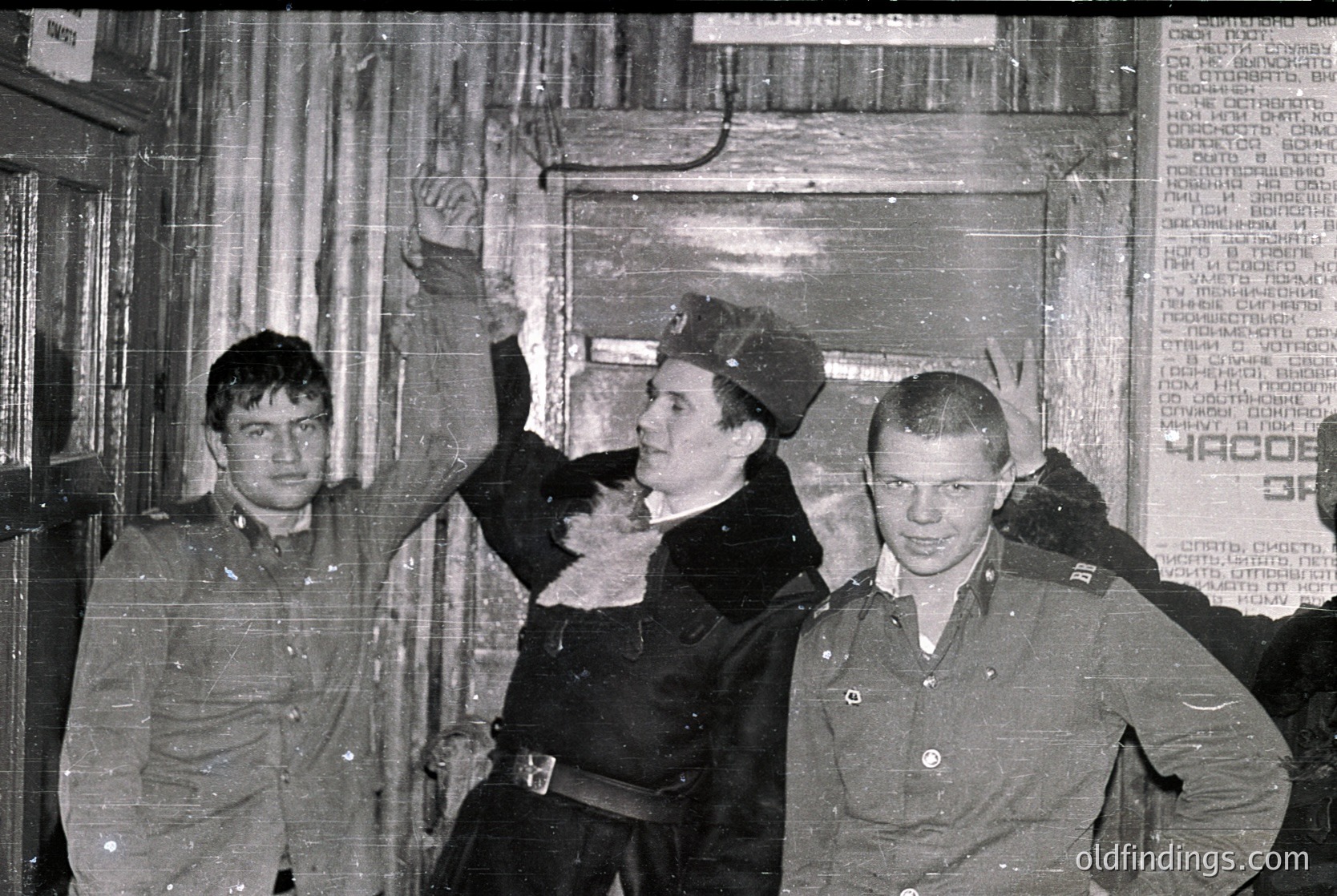 Three uniformed men pose indoors, likely Soviet-era military or state security personnel. The central figure gestures dramatically, while the others stand in a relaxed stance. Posters with Cyrillic text and a partially visible sign reading "КРОВЬ" (Blood) suggest a Soviet or Eastern Bloc setting. The grainy, monochrome style indicates a mid-20th-century photograph, likely 1960s–1980s.