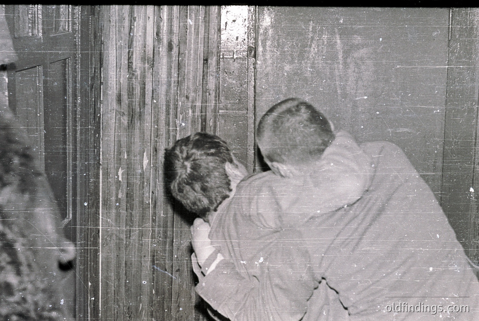 Two individuals in mid-20th century clothing examine a weathered, vertically striped wall—likely industrial or institutional. The man on the right wears a light jacket, while the other’s face is obscured. The grainy, monochrome tone suggests a vintage or photograph.