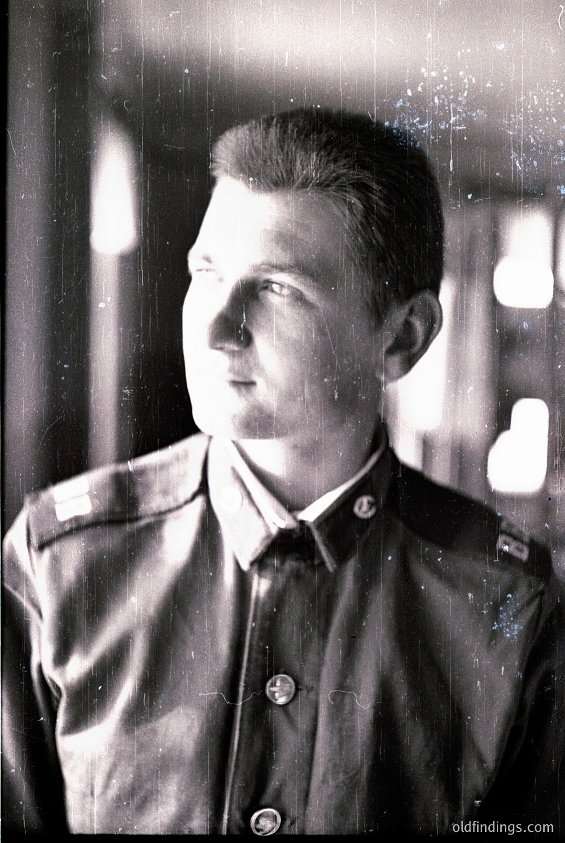 Portrait of a young man in a military-style uniform, likely from the mid-20th century. The uniform features shoulder boards and a collar insignia, suggesting a structured military or paramilitary organization. The setting appears to be indoors, possibly a studio, with a blurred background and light source behind him. The image style is black-and-white, indicative of the era. [mid20thcenturymilitary ]
