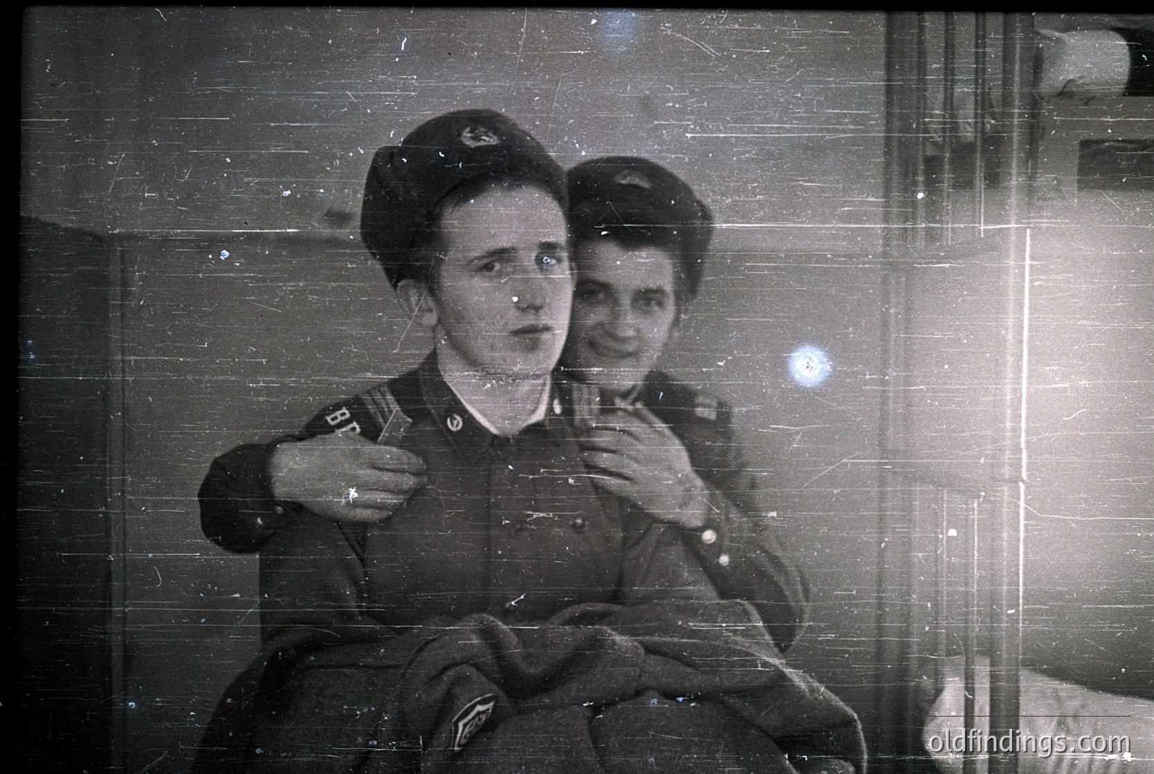 Vintage black-and-white photo of two individuals in military uniforms, likely Soviet-era (1940s–1950s). Uniforms feature peaked caps with insignia, suggesting military or paramilitary affiliation. One person holds a small object (possibly a cigarette or badge). Indoor setting with blurred background, indicating a train or confined space. Authentic historical documentation of mid-20th-century Soviet life.