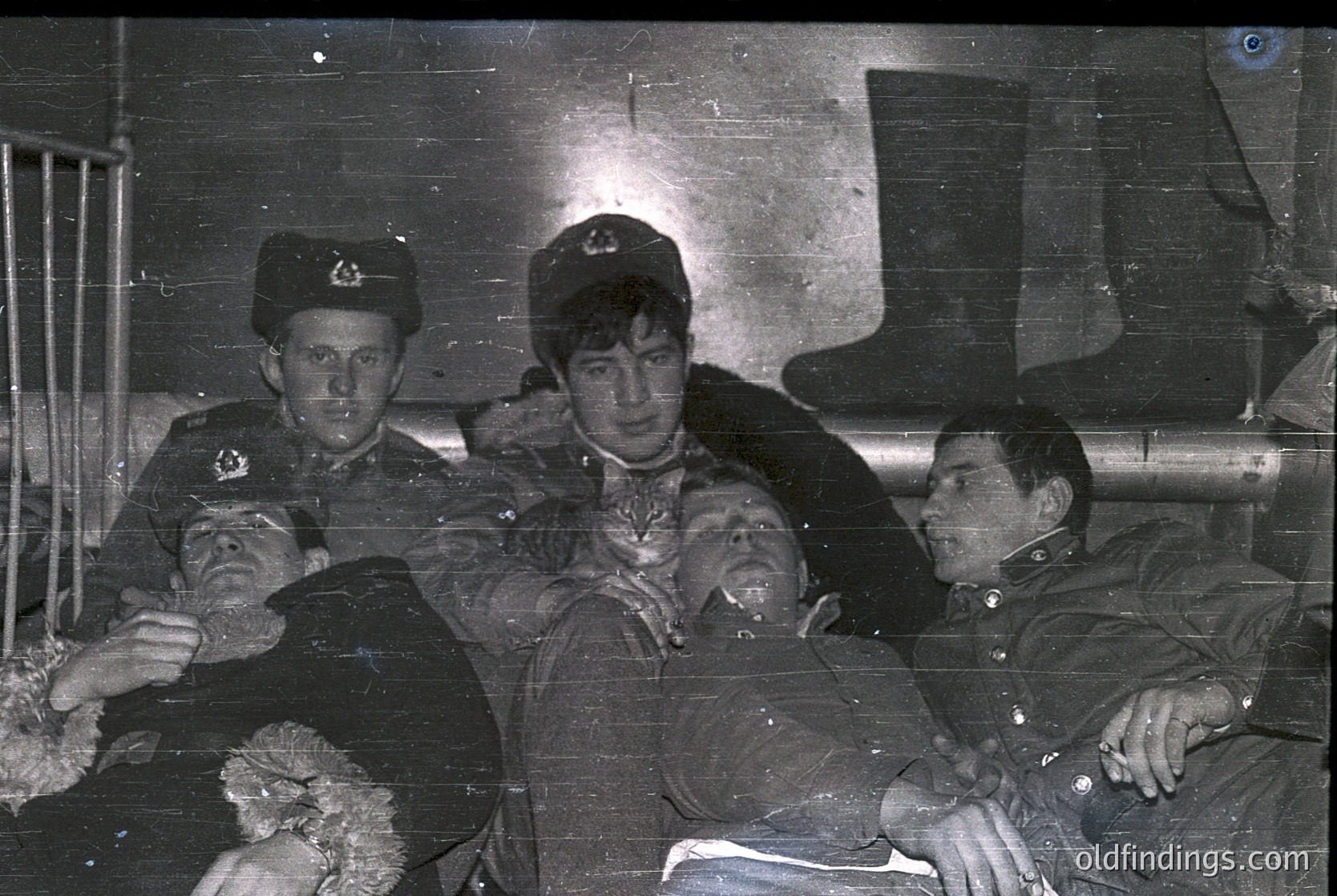 Vintage black-and-white photo of three uniformed men in a dimly lit, industrial setting—likely a train or ship cabin. Left: soldier in peaked cap with insignia. Center: seated man in formal attire, possibly military or official. Right: seated man in dark uniform, hand resting on knee. Visible pipes and metal railings suggest mid-20th century transport.