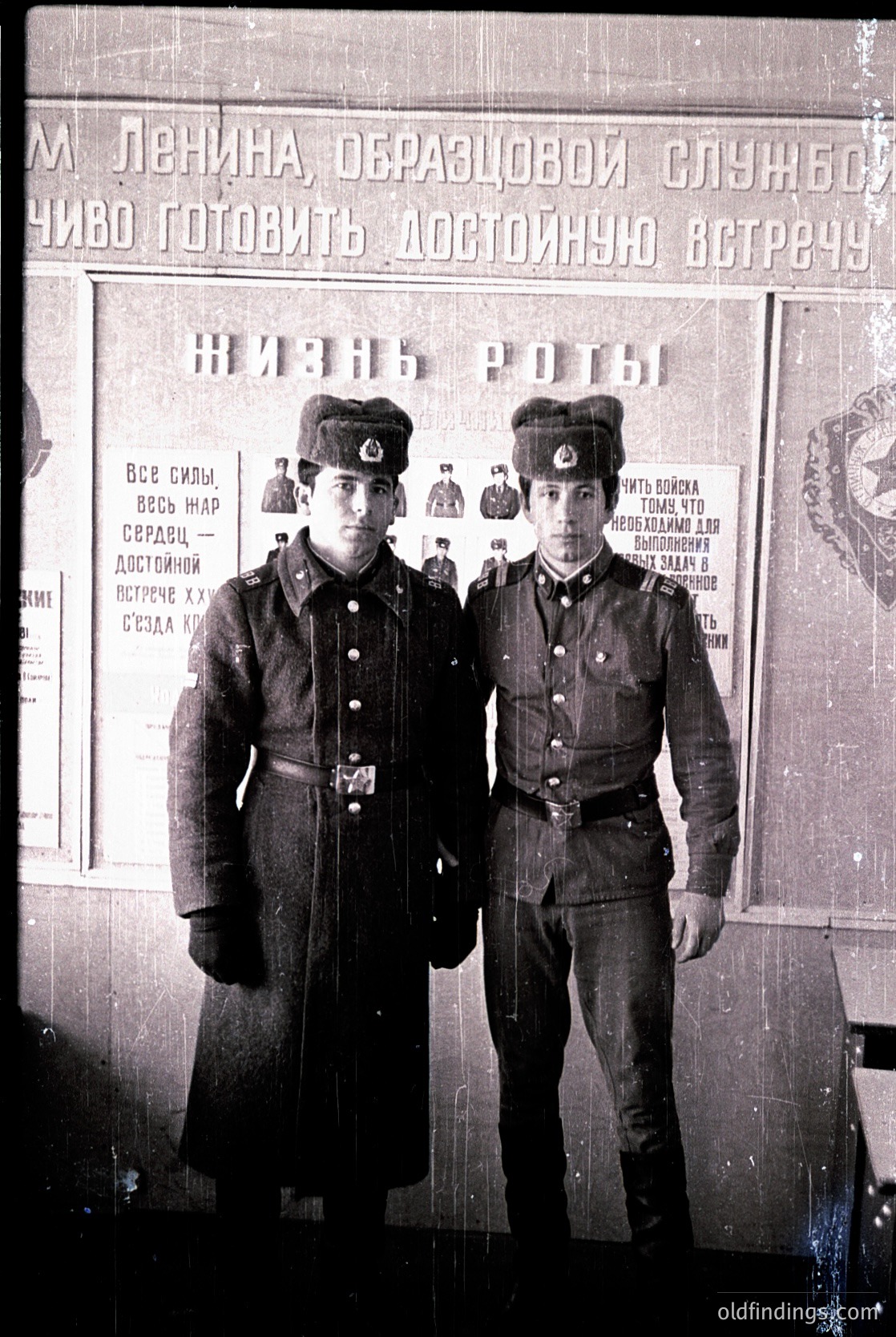 Vintage black-and-white photo of two young men in Soviet-era military uniforms, posed formally against a propaganda poster. Text in Cyrillic references Lenin and military service ideals. Uniforms include peaked caps with red stars, overcoats, and belts. Likely USSR, 1920s–1940s.