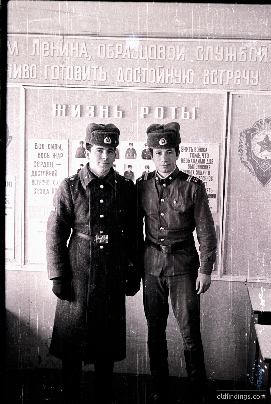 Two young men in Soviet-era military uniforms pose indoors, 1940s–1950s. Uniforms feature red star insignias, peaked caps, and belted coats. Poster in background reads Cyrillic slogans about Lenin and military service. Likely USSR or Eastern Bloc.
