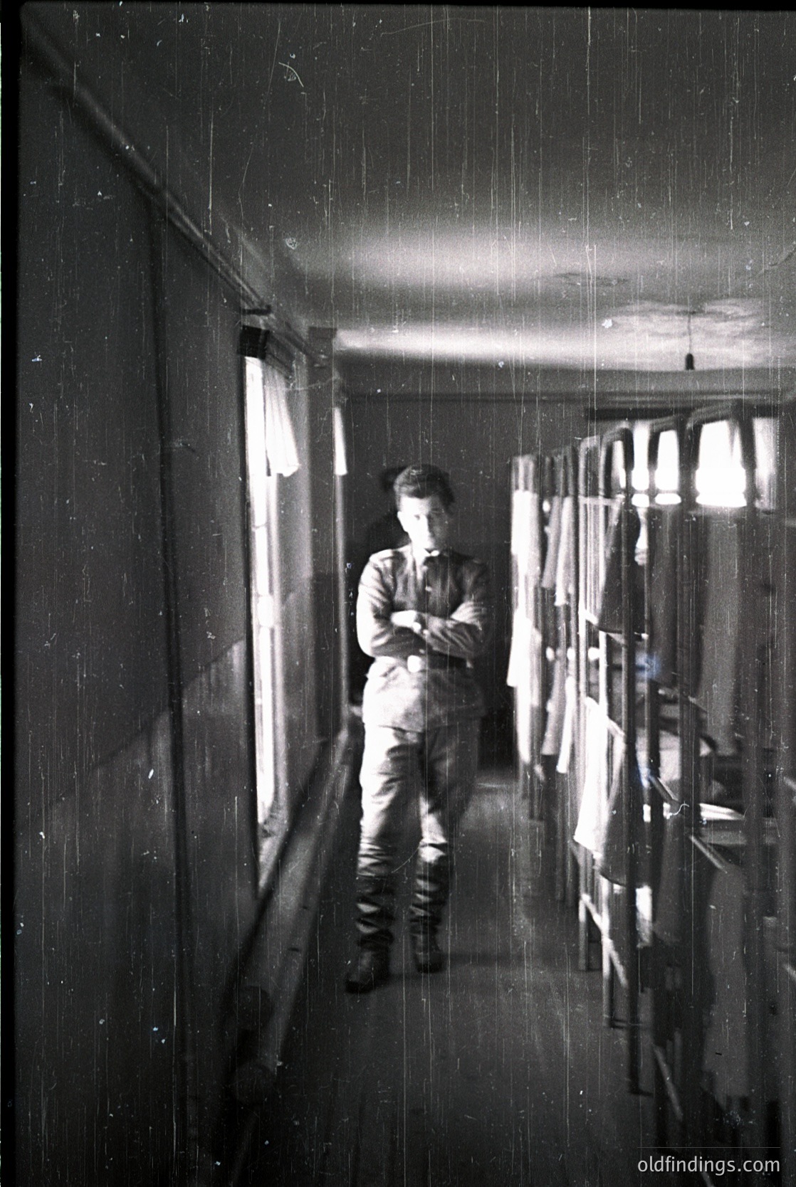 Mid-20th century industrial corridor with worker in overalls, standing near open doorway. Exposed pipes, bare lightbulbs, and utilitarian shelving reflect dim lighting.