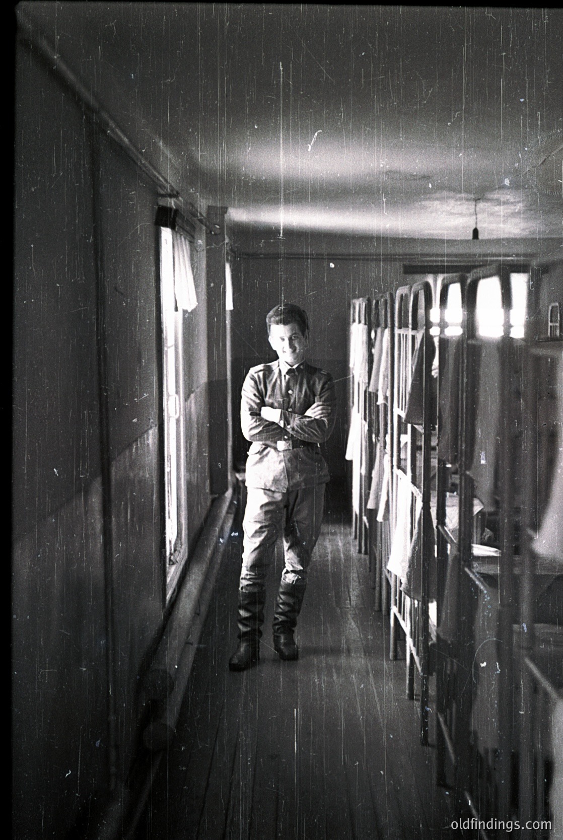 Mid-20th century man in uniform stands in a dimly lit, narrow hallway with rain streaks on windows. Industrial or institutional setting, likely a factory or warehouse. Uniform suggests military or labor discipline ( ).
