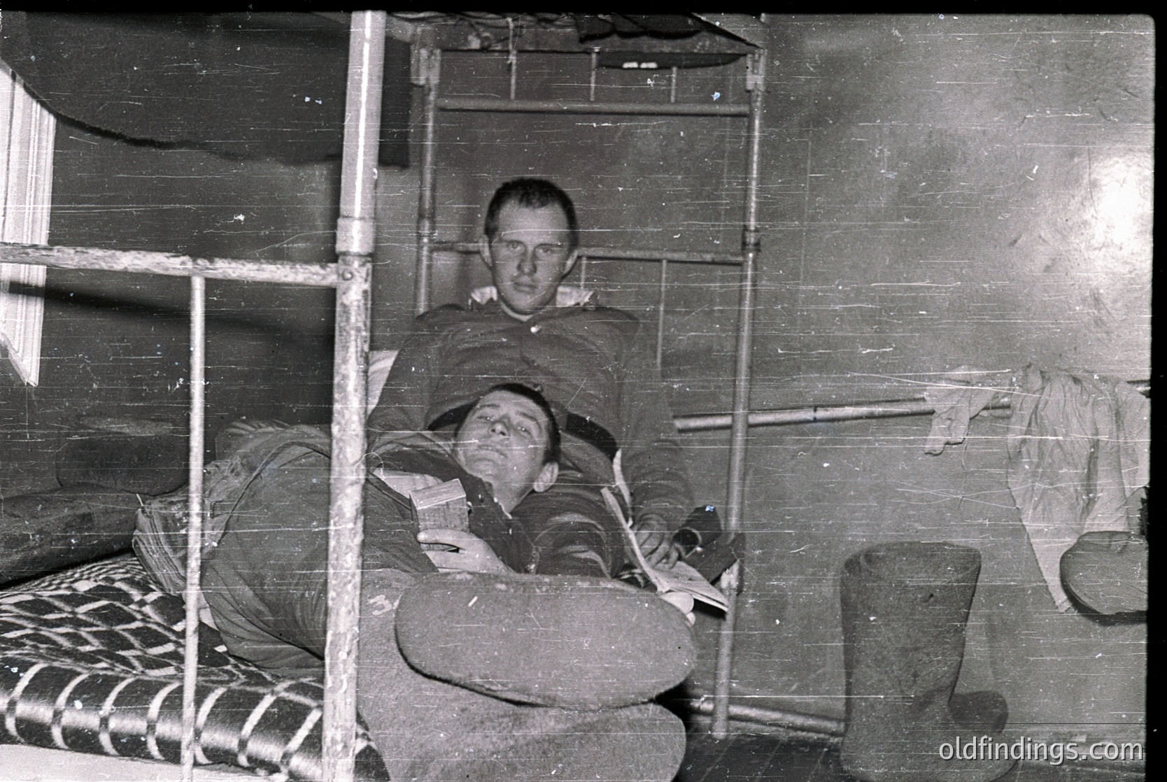 Two individuals seated on bunk beds in a confined, utilitarian space, likely a barracks or dormitory. The man on top holds a book; the lower bunk has a striped blanket and a pillow. Metal bars and a rifle rack suggest military or institutional use. Black-and-white, suggesting mid-20th century (1940s–1960s). [Two soldiers resting on bunk beds in a military barracks, mid-20th century ]