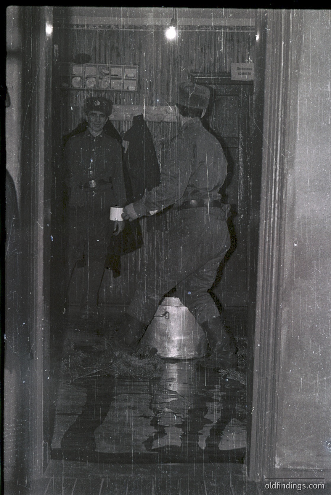 Two uniformed soldiers in heavy winter gear stand in a flooded subway entrance, 1960s-70s Eastern Bloc. One holds a helmet, the other a shovel. Reflections reveal a chaotic, waterlogged station with visible tracks and signage.
