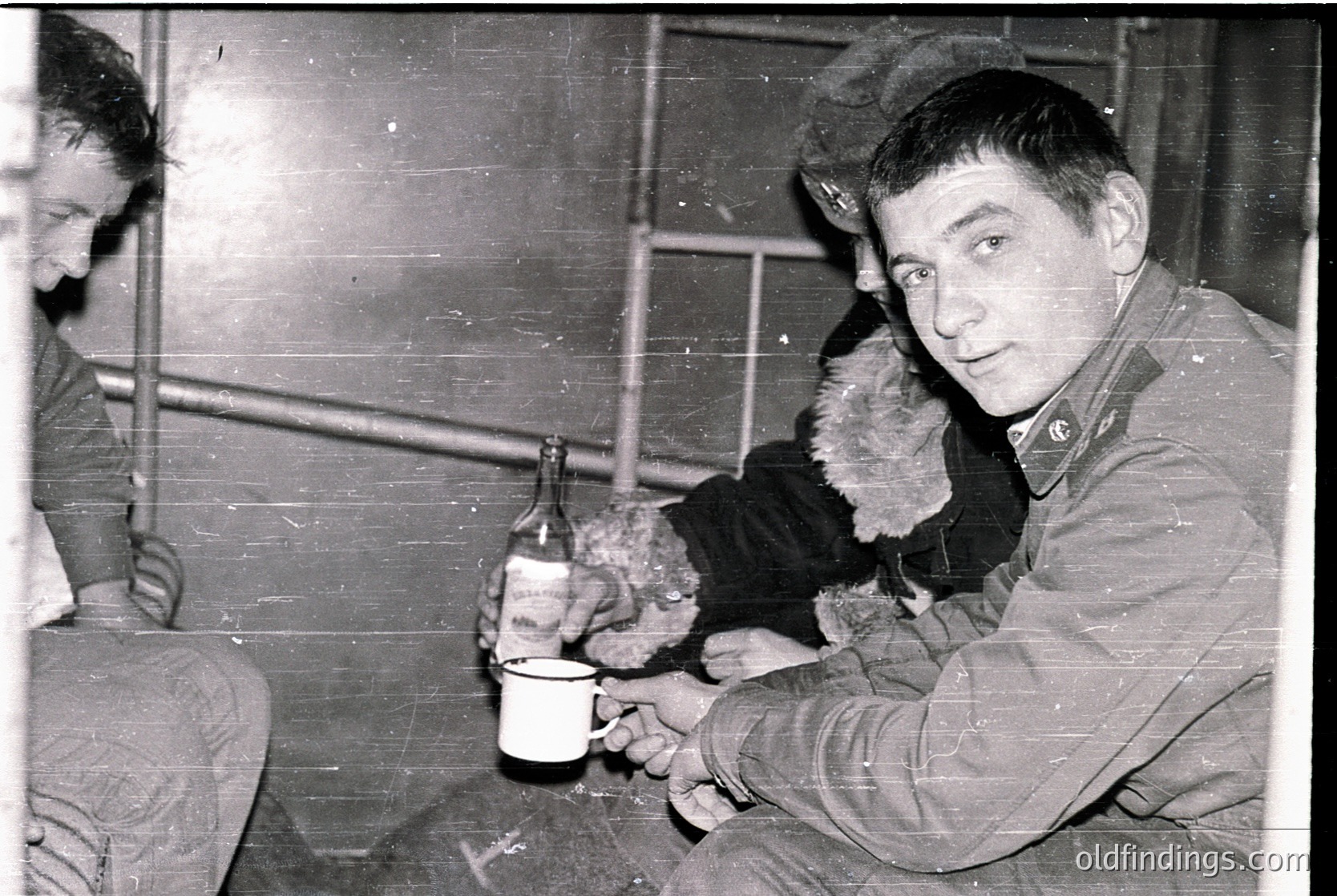 Black-and-white photo of two men in a dimly lit, industrial setting—likely a factory or warehouse. One man holds a bottle and cup, while the other sits in the background. Heavy winter coats and fur-lined hoods suggest cold weather, possibly 1950s–1970s Eastern Europe. Rustic brick walls and metal scaffolding add to the utilitarian atmosphere.