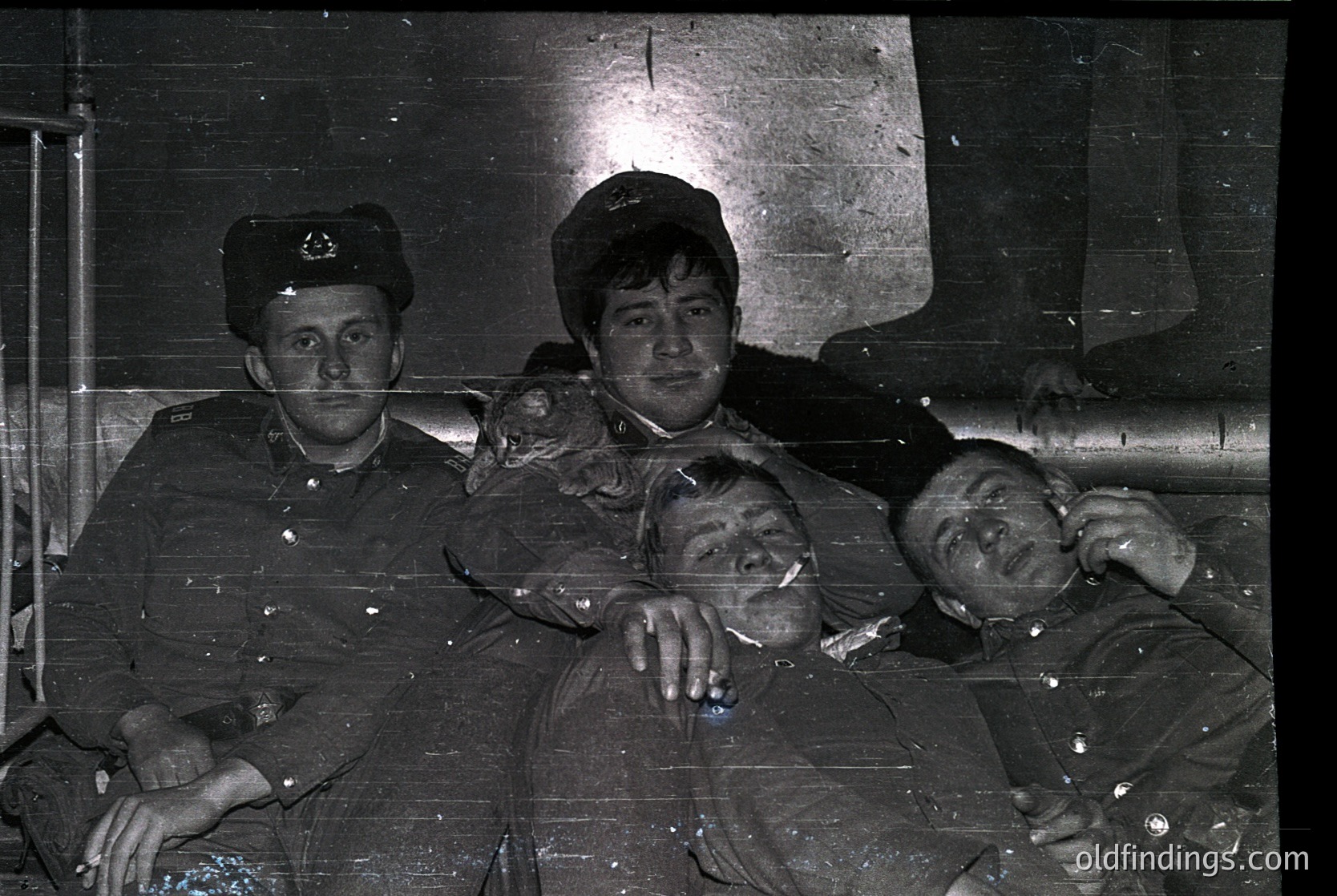 Three young men pose indoors in Soviet-era military uniforms, likely late 1940s–1960s. Uniforms feature peaked caps with insignia, suggesting naval or air force affiliation. Dark, utilitarian setting with exposed pipes and concrete walls. Candid expressions and relaxed postures hint at camaraderie.