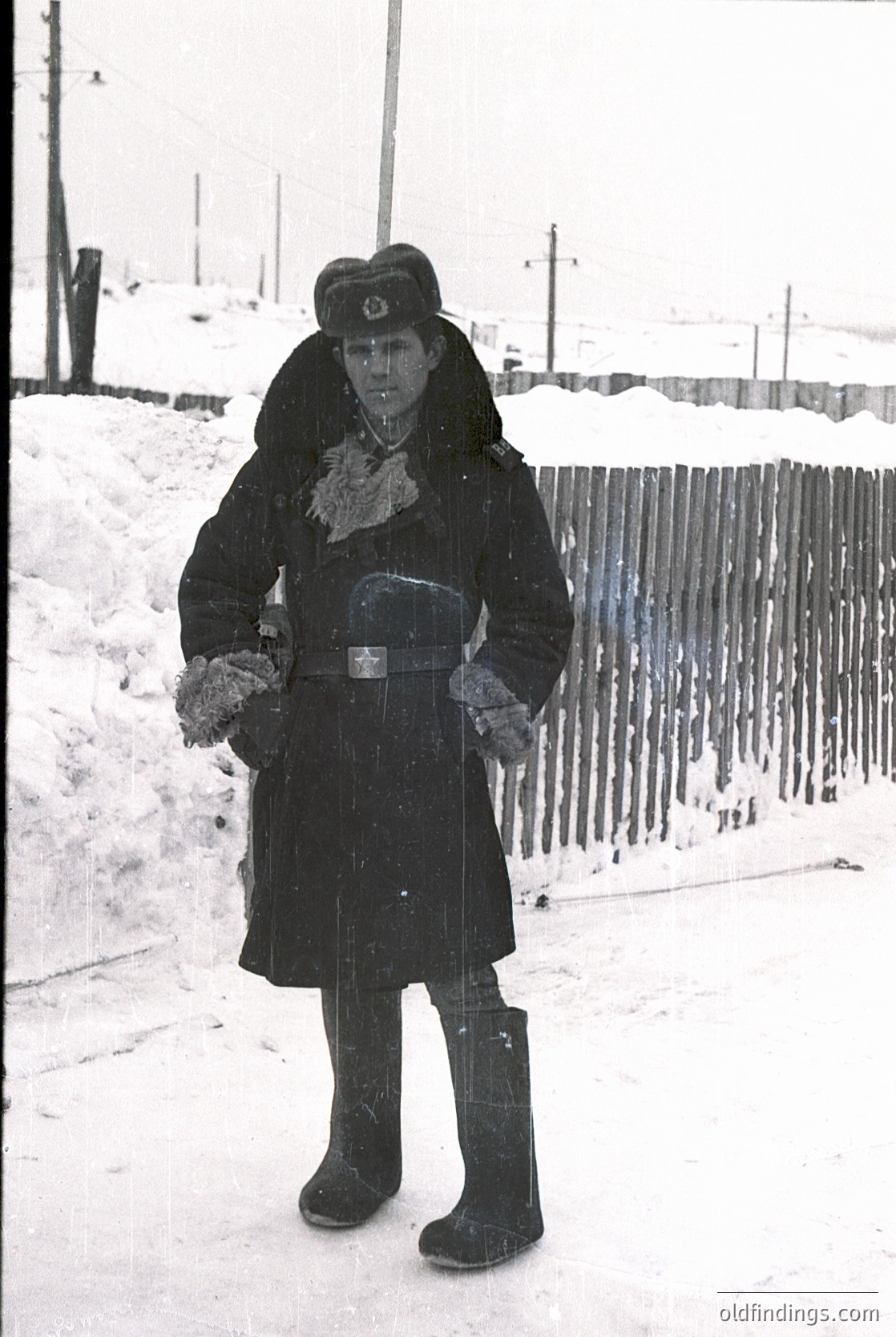 Winter uniform with fur collar and belt, holding mittens. Snow-covered fence and ground. Mid-20th century Soviet-era attire.