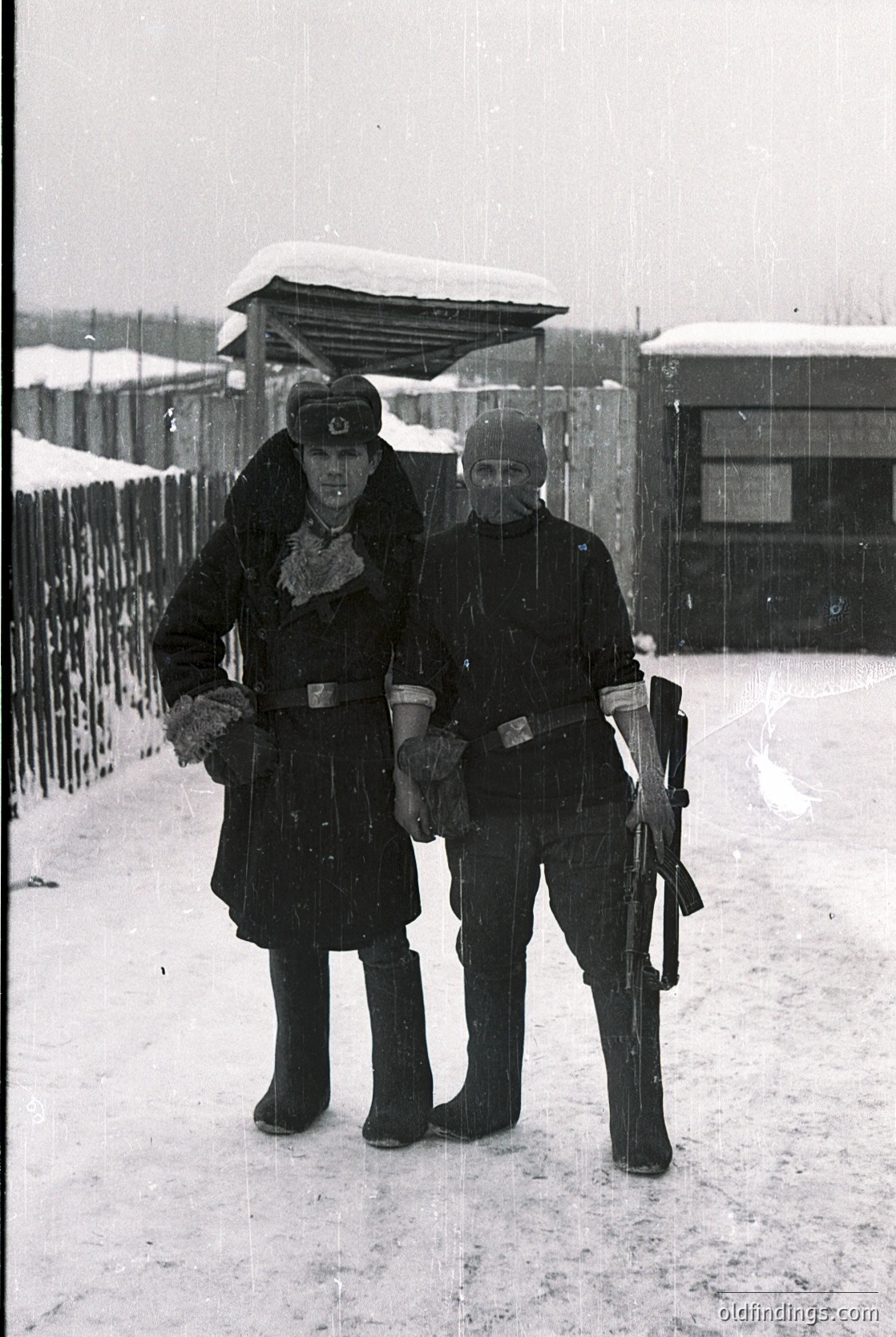 Two uniformed soldiers pose in winter attire, likely WWII-era German Wehrmacht. Snow-covered ground and barbed wire fence suggest a guarded area. Distinctive caps, belts, and rifles indicate military discipline.