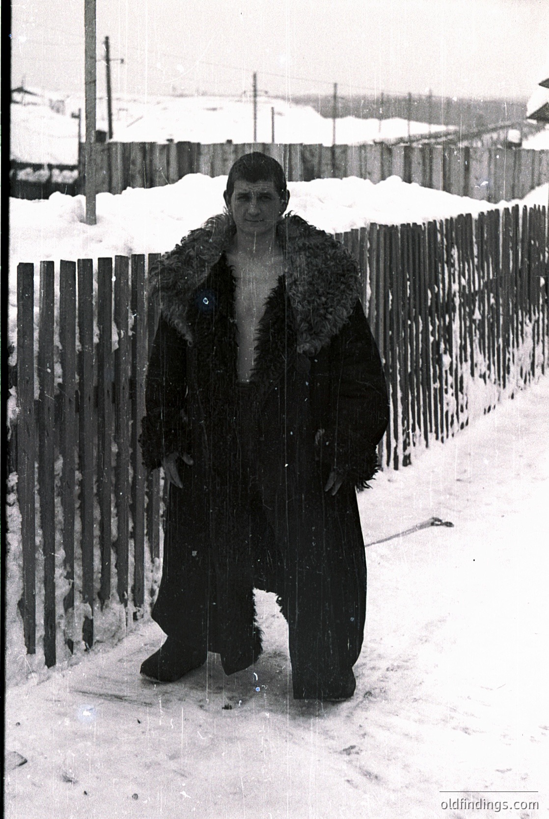 Portrait of a man in a heavy fur-lined coat, standing in snow-covered urban setting. Wooden fence and residential buildings in background suggest mid-20th century Eastern European winter. Coat style hints at 1950s-1960s practical winterwear.