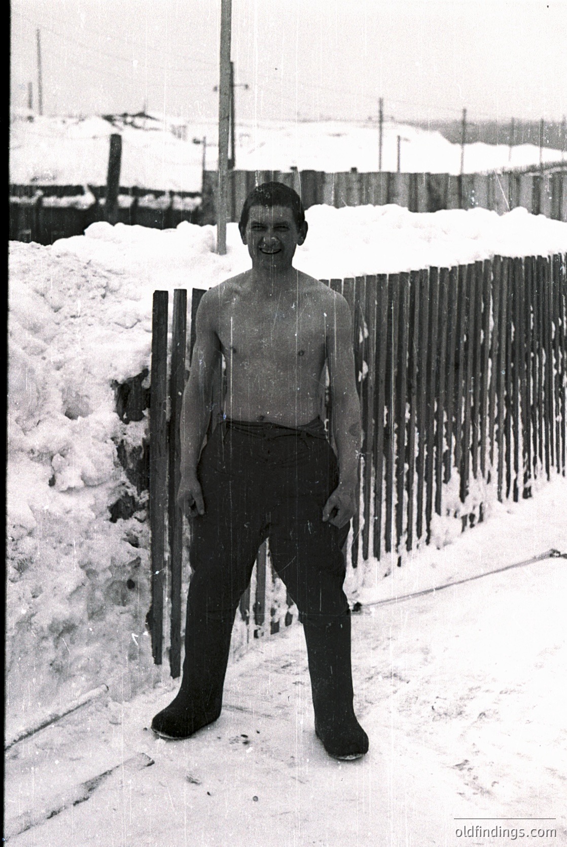 Young boy in mid-20th-century winter attire—shirtless with long wool socks and heavy boots—poses beside a snow-laden wooden fence. Snowdrifts and industrial buildings in background suggest urban setting. Likely or .