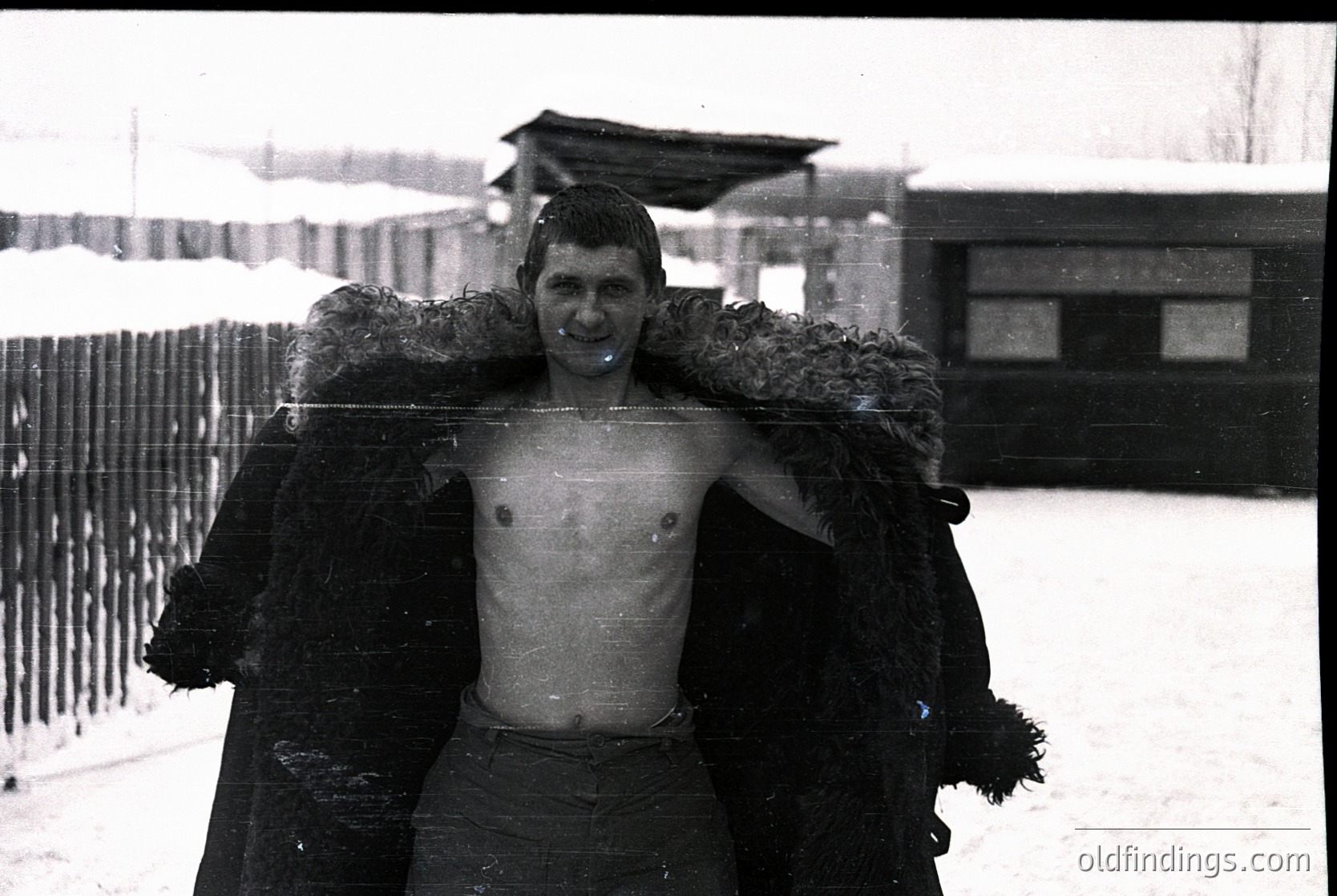 Young man in mid-20th century winter attire, draped in a fur-lined coat while shirtless, standing in snow-covered surroundings. Snowflakes cling to his shoulders and coat. Background shows a fence, bare trees, and a building—likely Eastern European or Soviet-era setting.