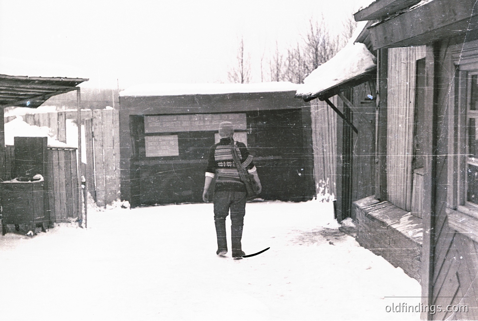 A man in a winter uniform, likely a postal worker, stands in a snowy alleyway holding mail. The scene features rustic wooden buildings with snow-covered roofs and a sign on a dark wooden door.