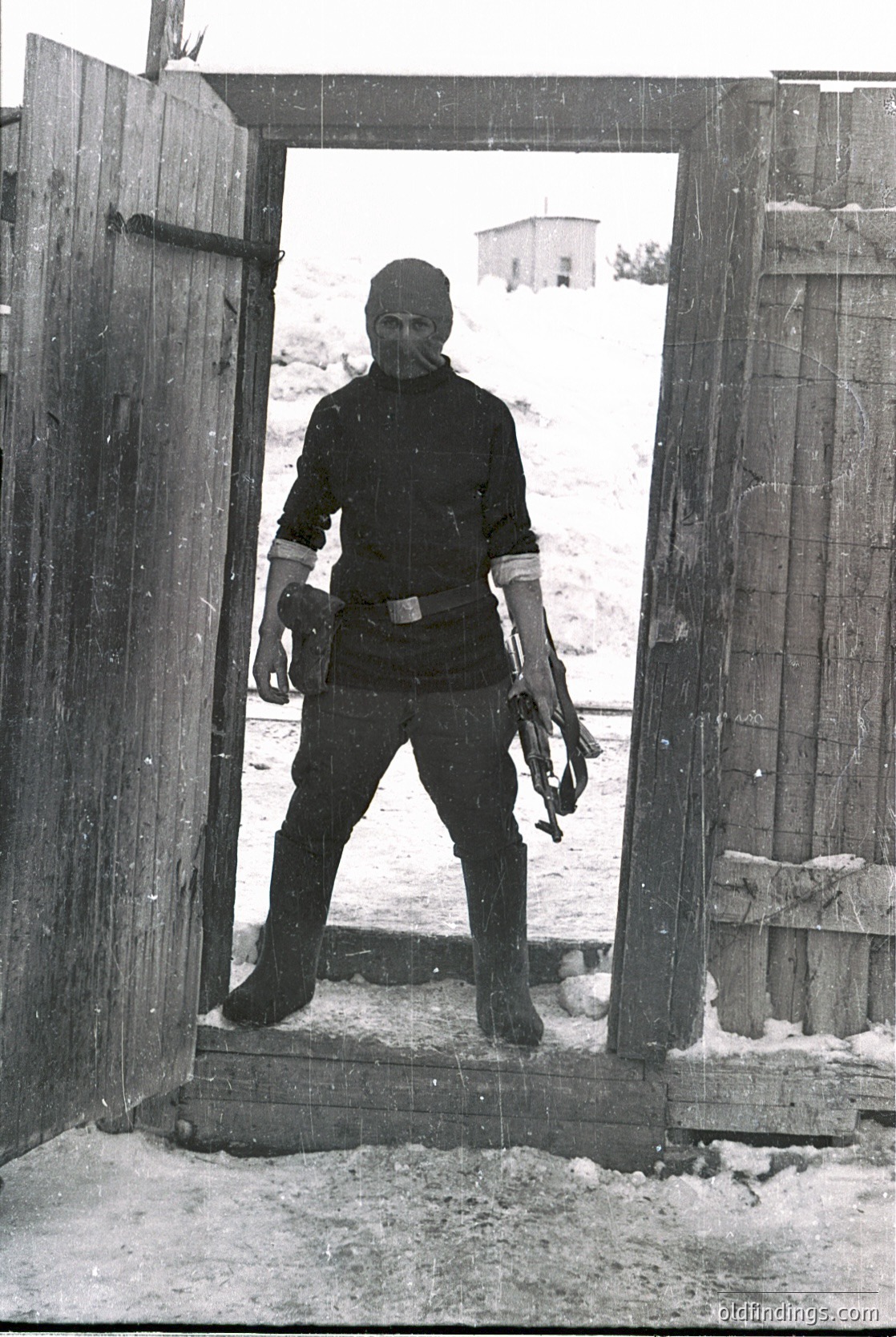 Cold-weather gear: heavy insulated coat, gloves, and ski goggles. Standing in a wooden archway, likely a ski lift entrance or chalet doorway. Snow-covered ground and rustic wooden structures in background. Mid-20th century alpine setting.