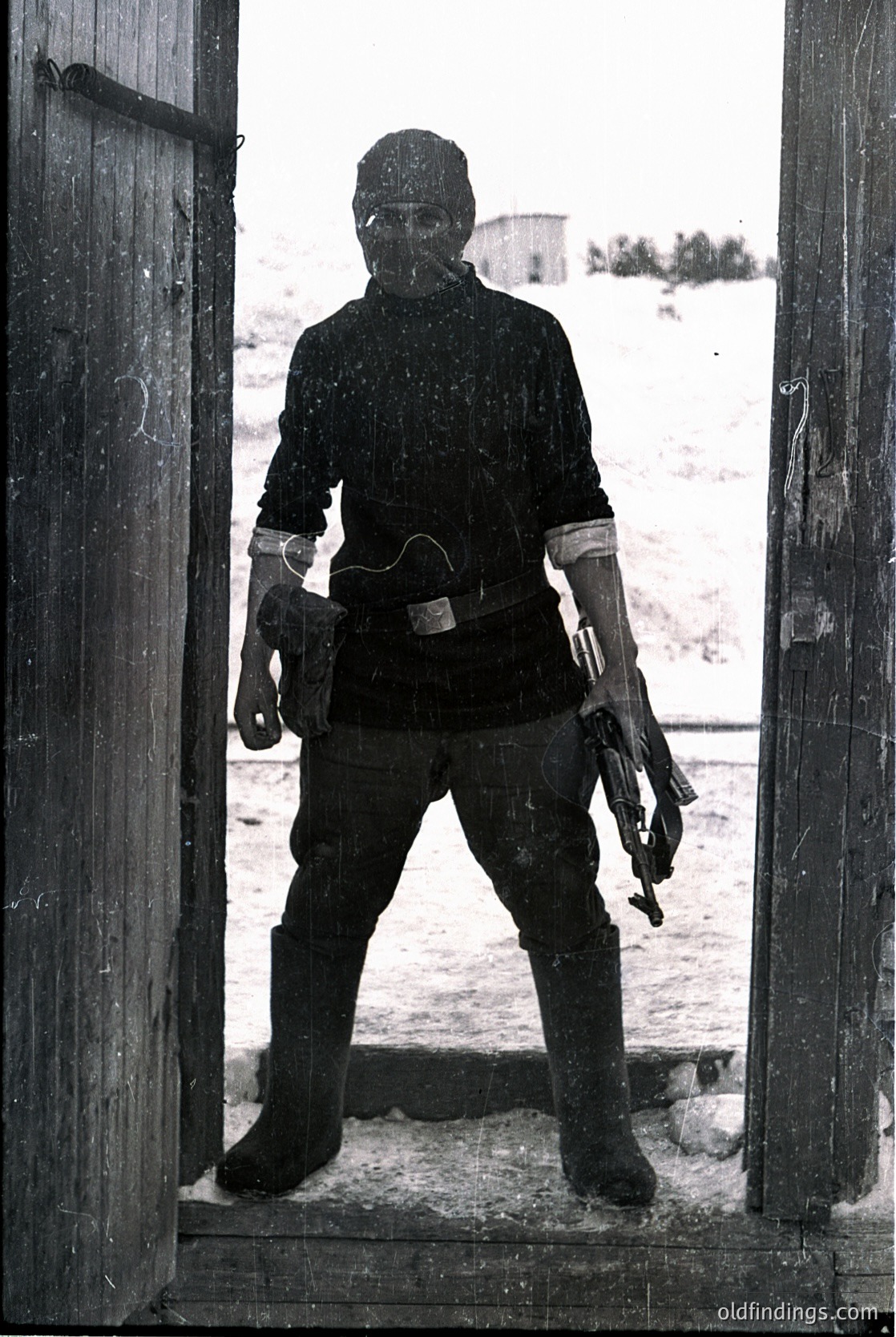Cold War-era soldier in full winter gear stands in doorway, gripping submachine gun. Snowy landscape and concrete structures suggest Eastern Bloc training facility, likely 1960s–1980s. Uniform includes padded jacket, gloves, and ski mask for Arctic conditions.