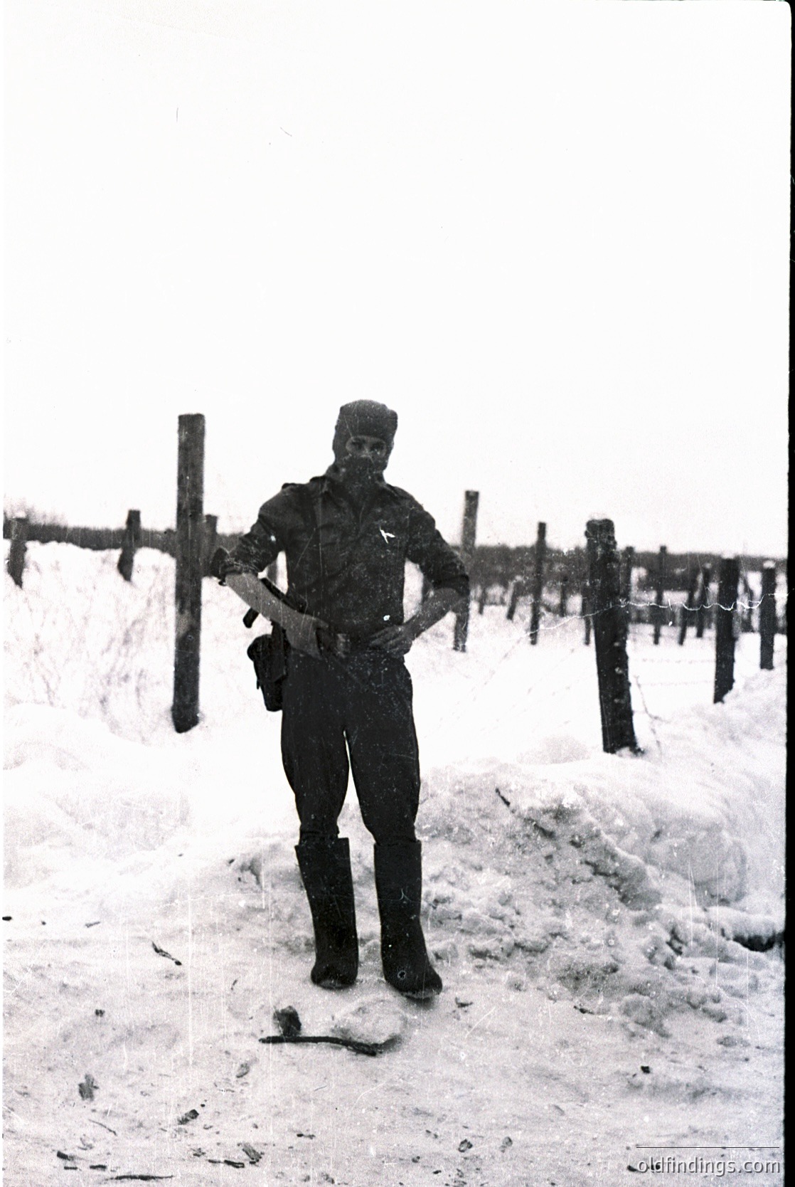 Soldier in winter gear poses by barbed-wire fence in snowy terrain, mid-20th century. Uniform suggests military training or guard duty.