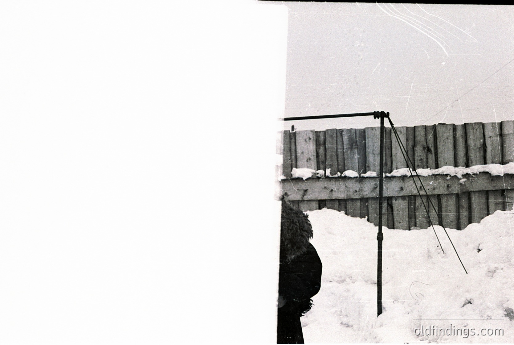 Black-and-white photograph of a rustic wooden fence with a slanted roof structure, likely a barn or storage shed. Snow covers the ground and fence base, indicating winter. The composition emphasizes texture and weathered wood grain. Potential agricultural or rural setting.