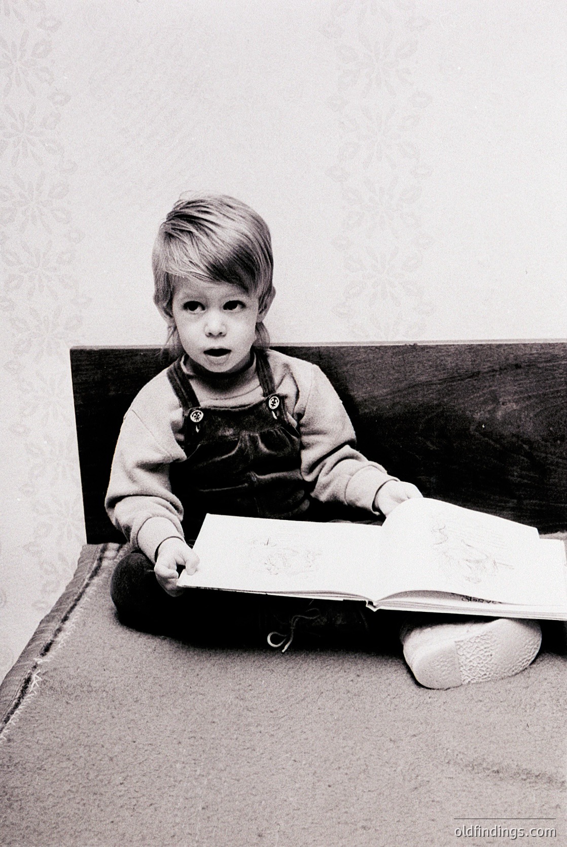 Young child in mid-20th-century overalls and slippers engrossed in an open book on a carpeted floor, seated beside a dark wooden bench. Classic mid-century domestic scene.