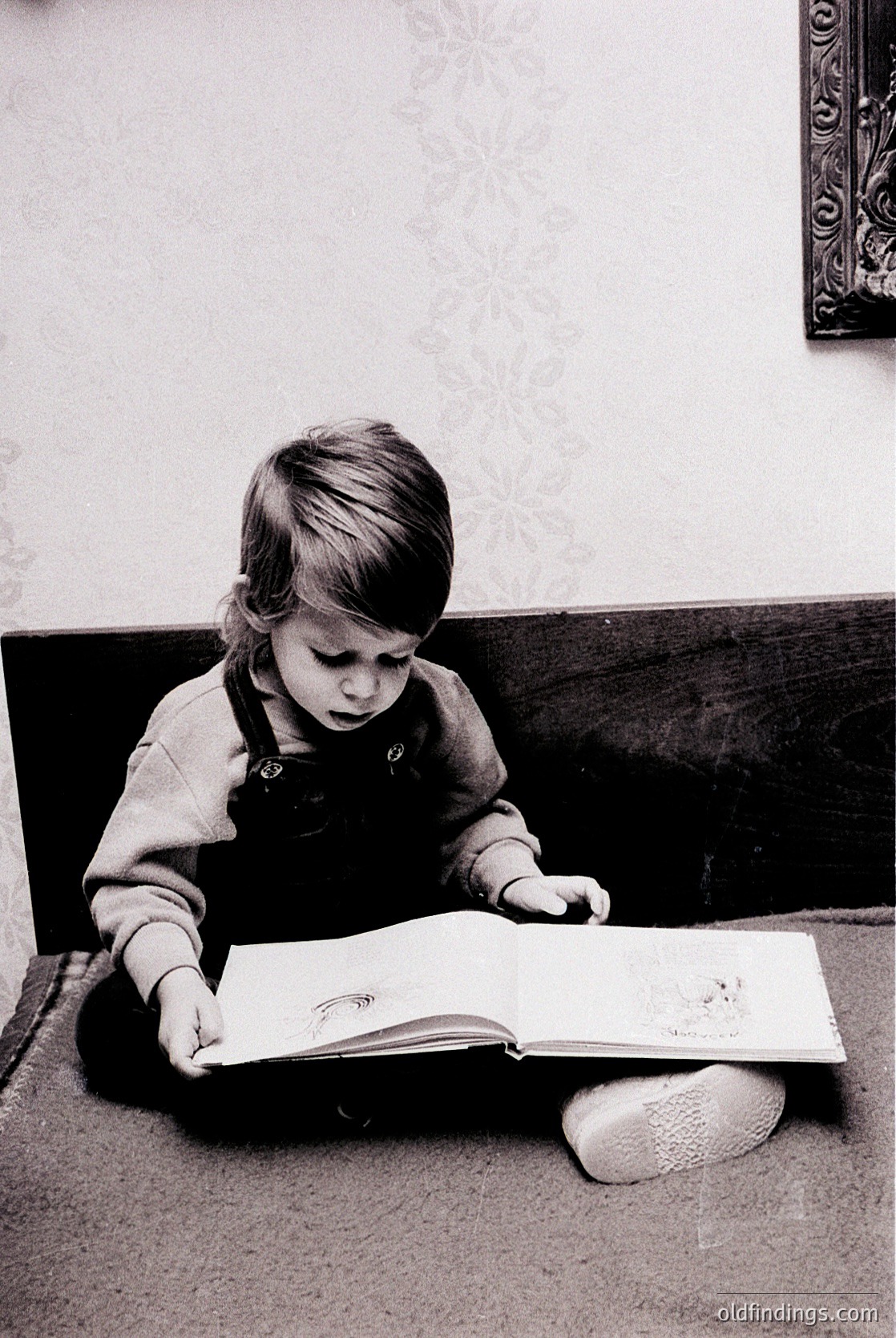 A young child engrossed in an open book on a dark carpeted floor, dressed in a long-sleeved shirt with suspenders and white slip-on shoes. The setting appears to be an indoor domestic space, likely mid-20th century. The child’s focused expression suggests early literacy engagement.