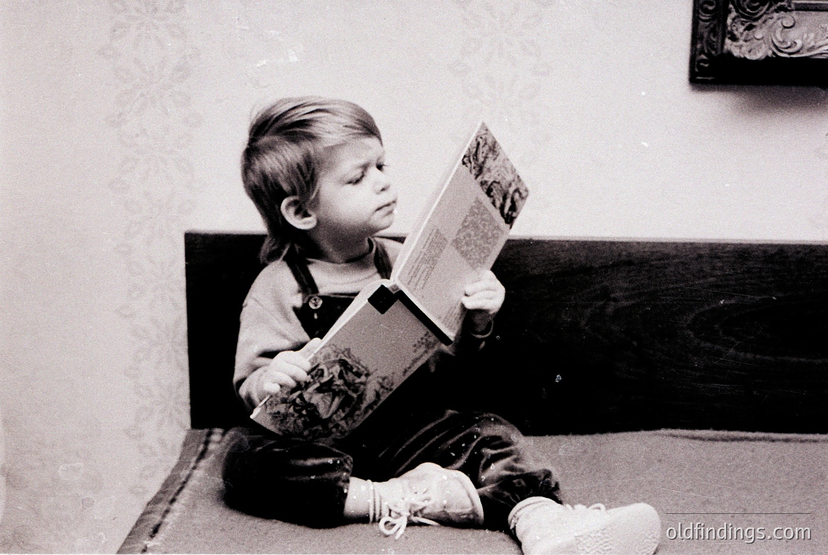 Black-and-white portrait of a young child engrossed in a large-format illustrated book, seated on a wooden bench. Overalls and white socks with tassels suggest mid-20th-century style. Plain wall and framed mirror in background indicate domestic setting.